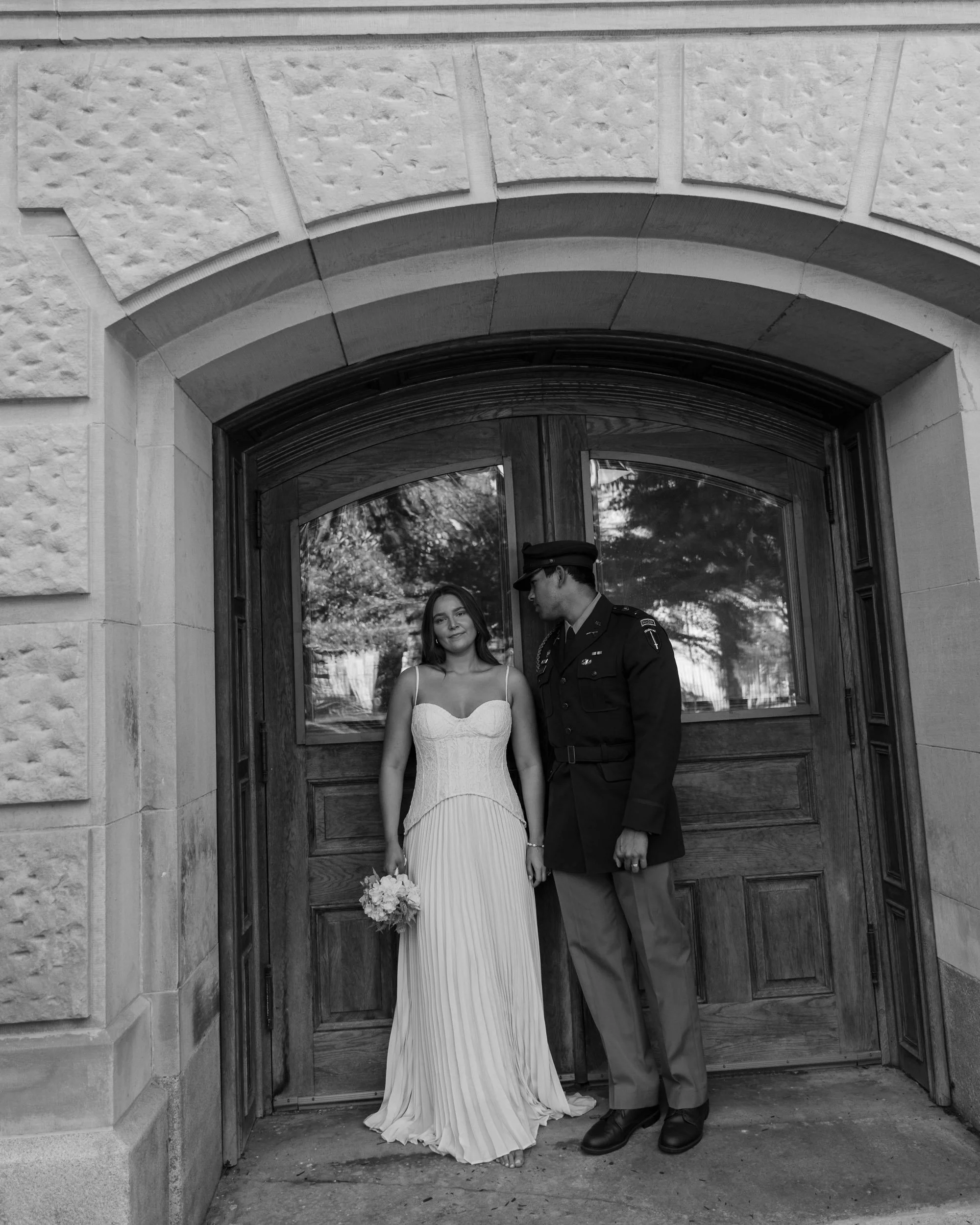 A woman in a wedding dress and a man in a military uniform standing under a stone archway with a wooden door and windows, outdoors.