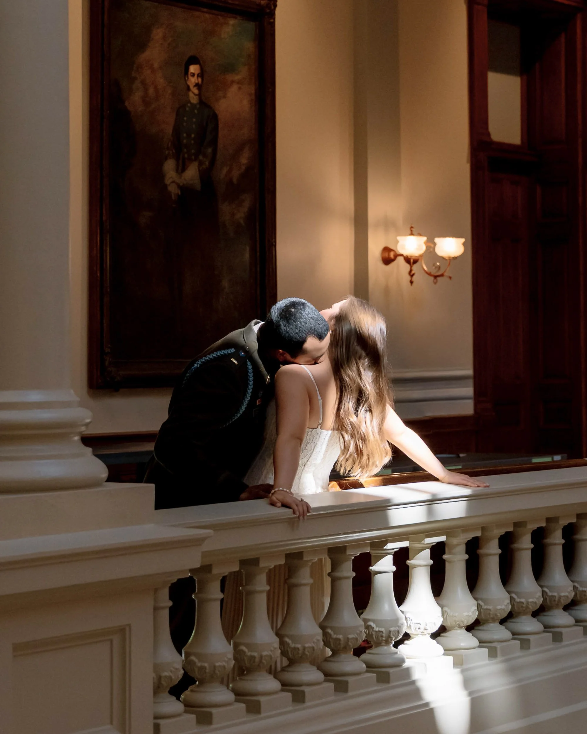 A man in a dark uniform and a woman in a white dress are sharing a kiss on a balcony inside a historic building, with a large portrait painting on the wall behind them.