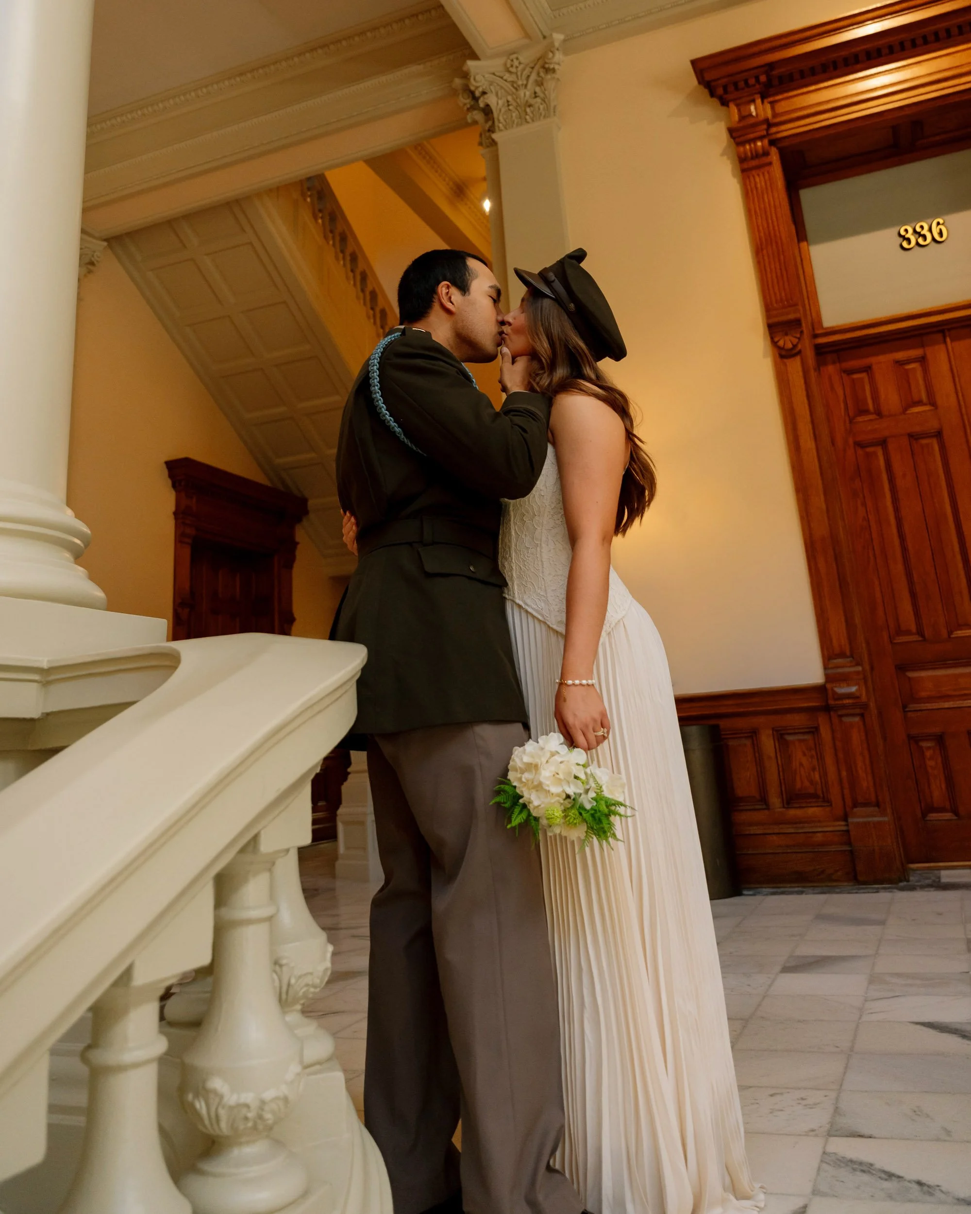A couple kissing inside a building with elegant architecture, the woman holding a bouquet of white flowers, wearing a white pleated dress and a black hat, and the man dressed in a military uniform.