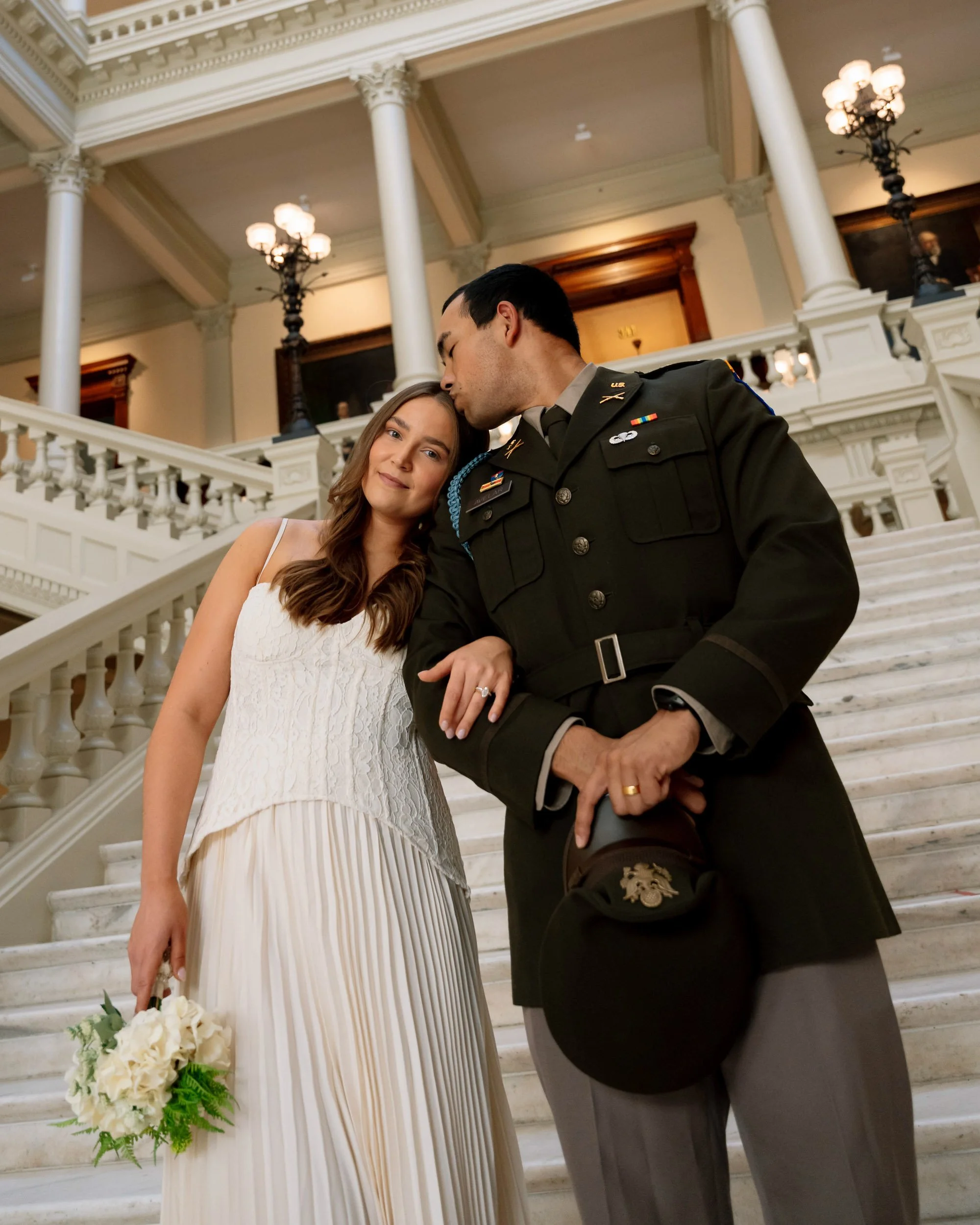 A woman in a white dress holding a bouquet, standing next to a man in a military uniform on a marble staircase inside a grand building, with ornate architectural details and decorative lighting.