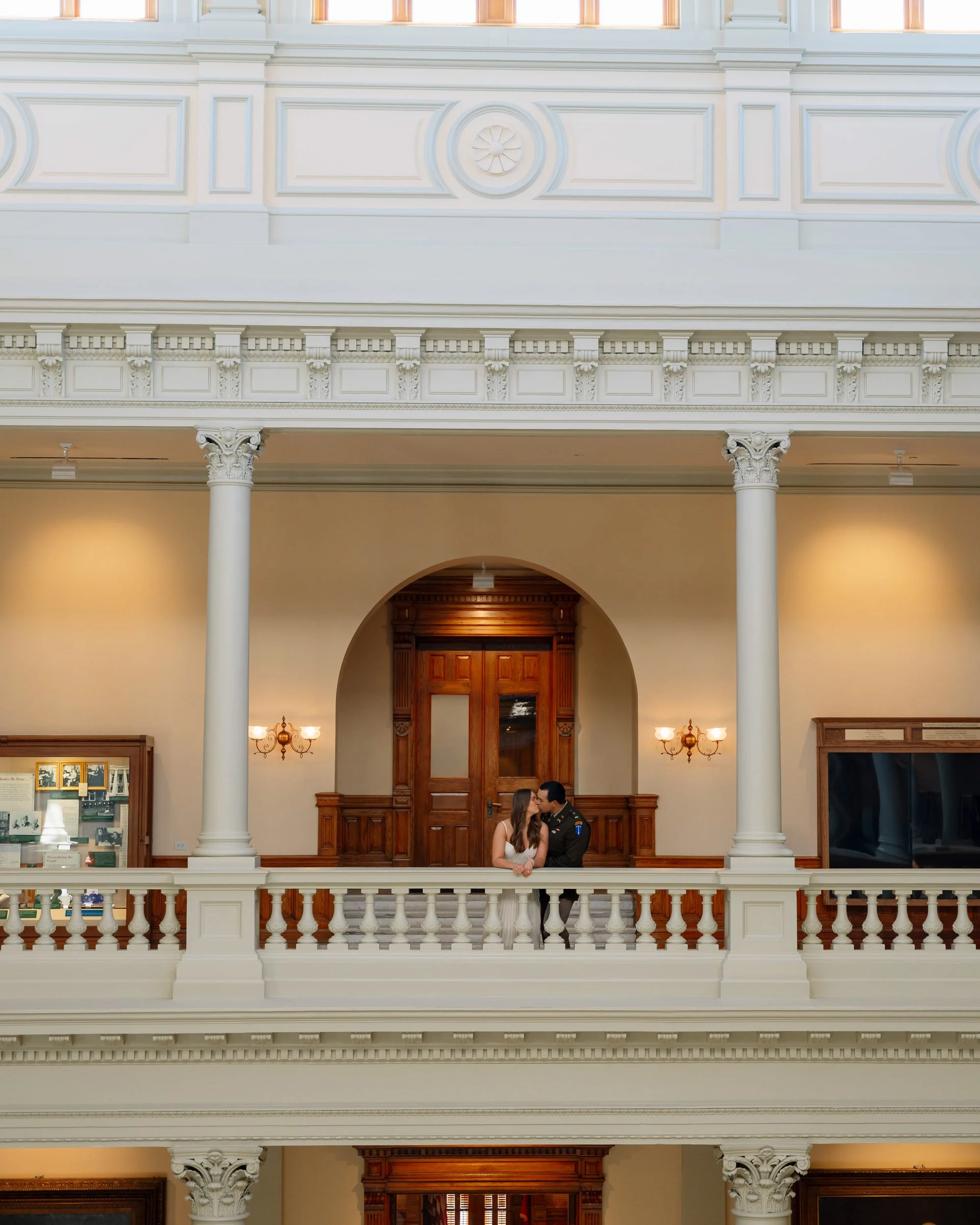 A young couple stands on an ornate balcony in a historic building, sharing a kiss. The interior features classical architecture with white columns, detailed moldings, and wooden doors.