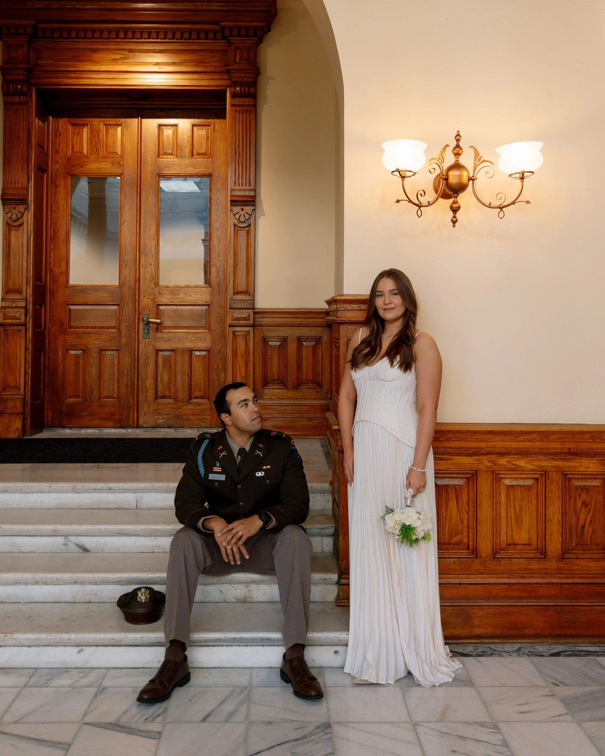 A man in military uniform sitting on marble stairs looking up at a woman in a white wedding dress holding a bouquet of white flowers, standing on the stairs in a wood-paneled hallway with a large wooden door and a vintage wall lamp.