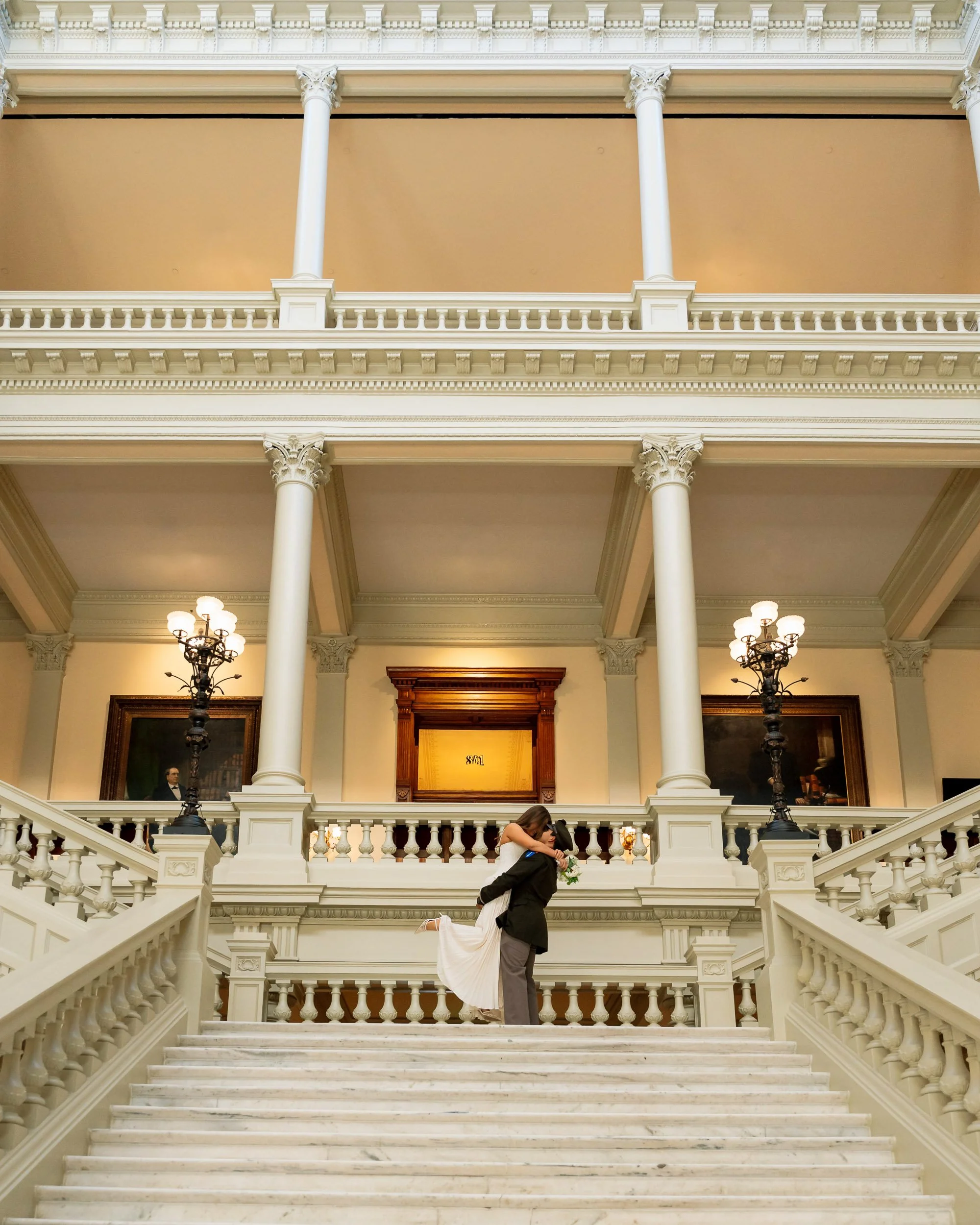 A couple dressed in wedding attire embracing on a grand staircase inside a historic building with ornate columns, chandeliers, and framed paintings.