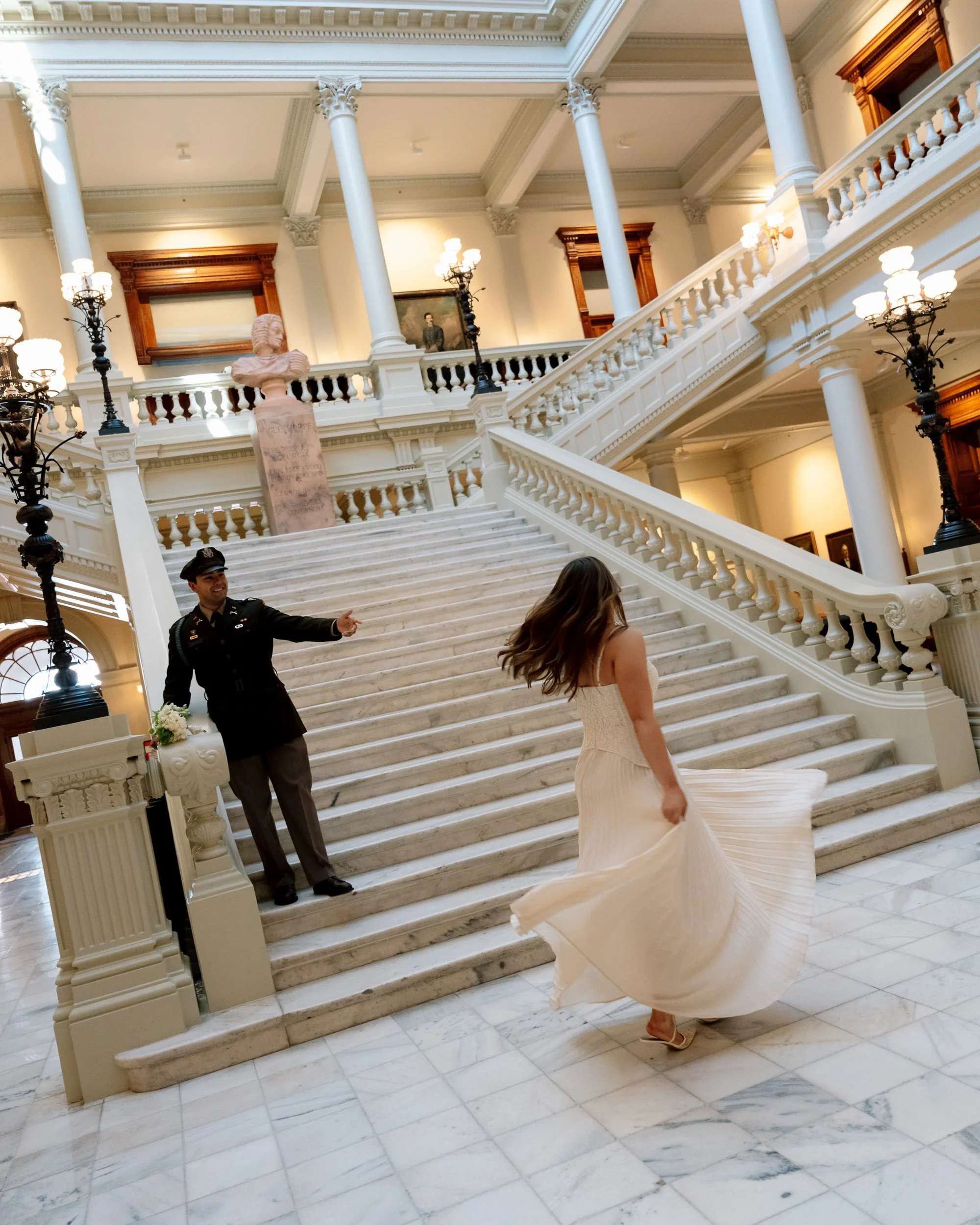 A woman in a white dress twirling on a marble staircase in a grand, historic building, while a man in a uniform points at her.
