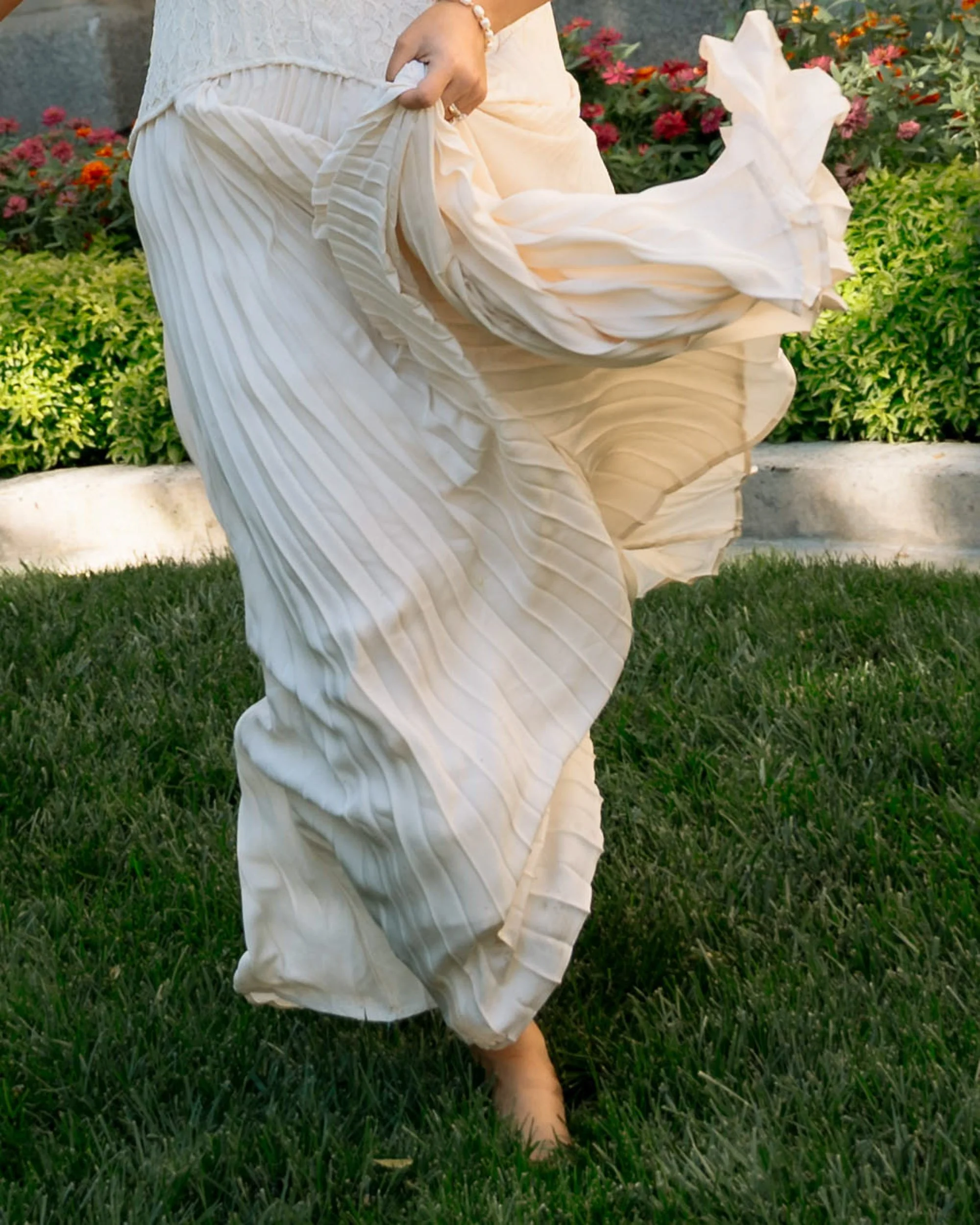 Close-up of a woman in a flowing white pleated dress, lifting the fabric with one hand while standing on grass in a garden with colorful flowers and green bushes in the background.
