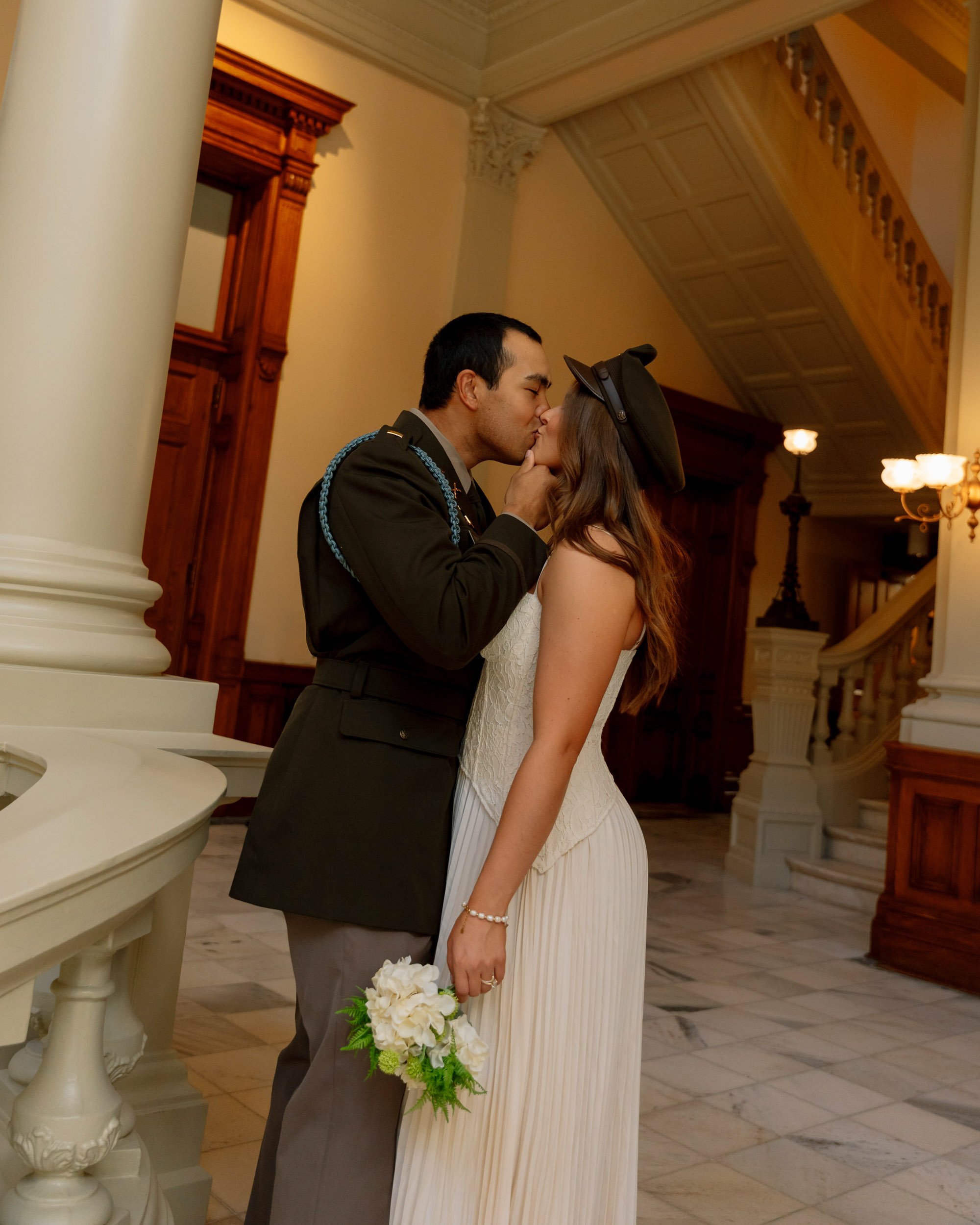 A couple sharing a kiss in formal attire inside a grand building with marble floors and wooden decor.