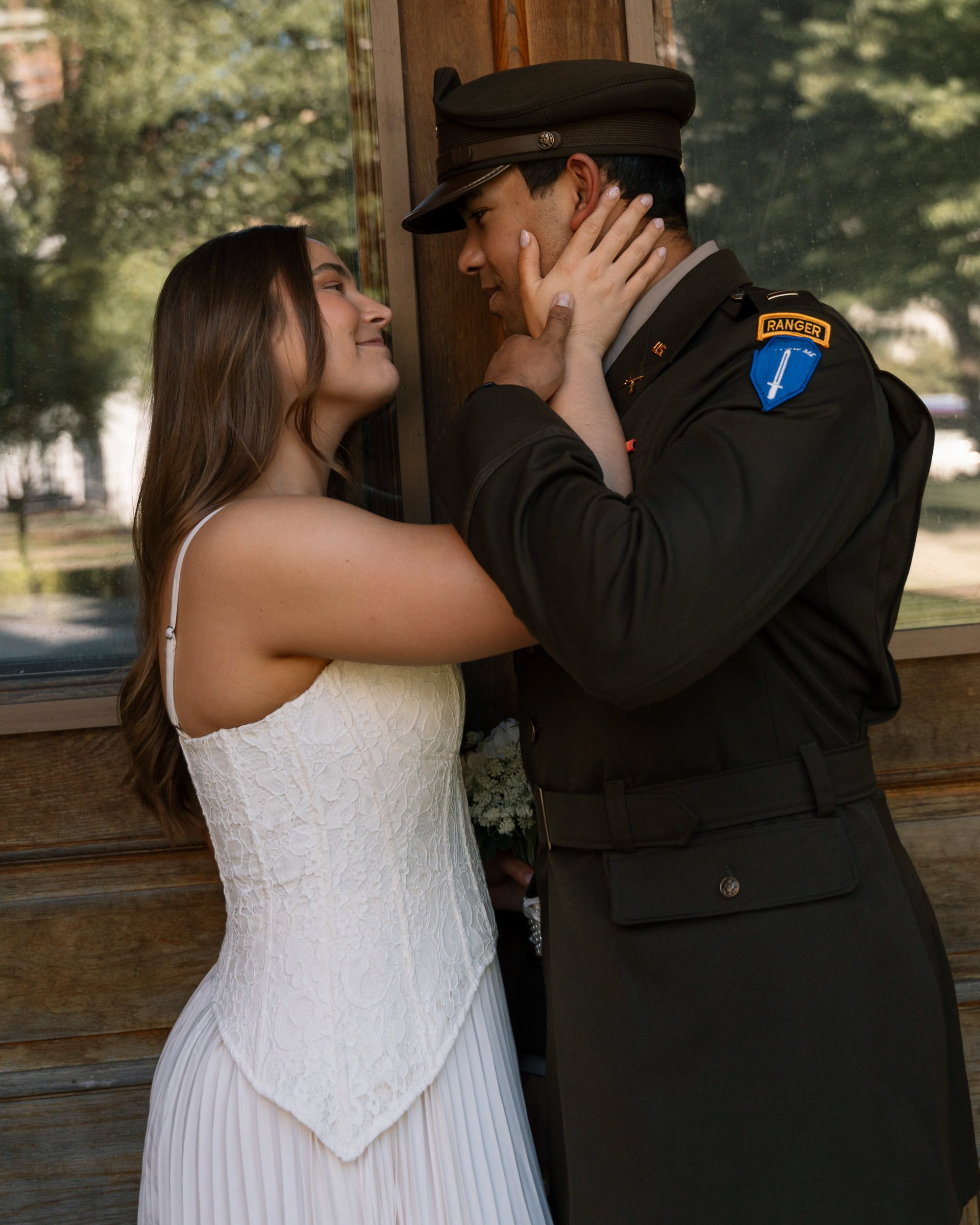 A young woman in a white dress and a man in a police uniform are standing close together, touching foreheads and smiling, in front of a wooden wall and window.