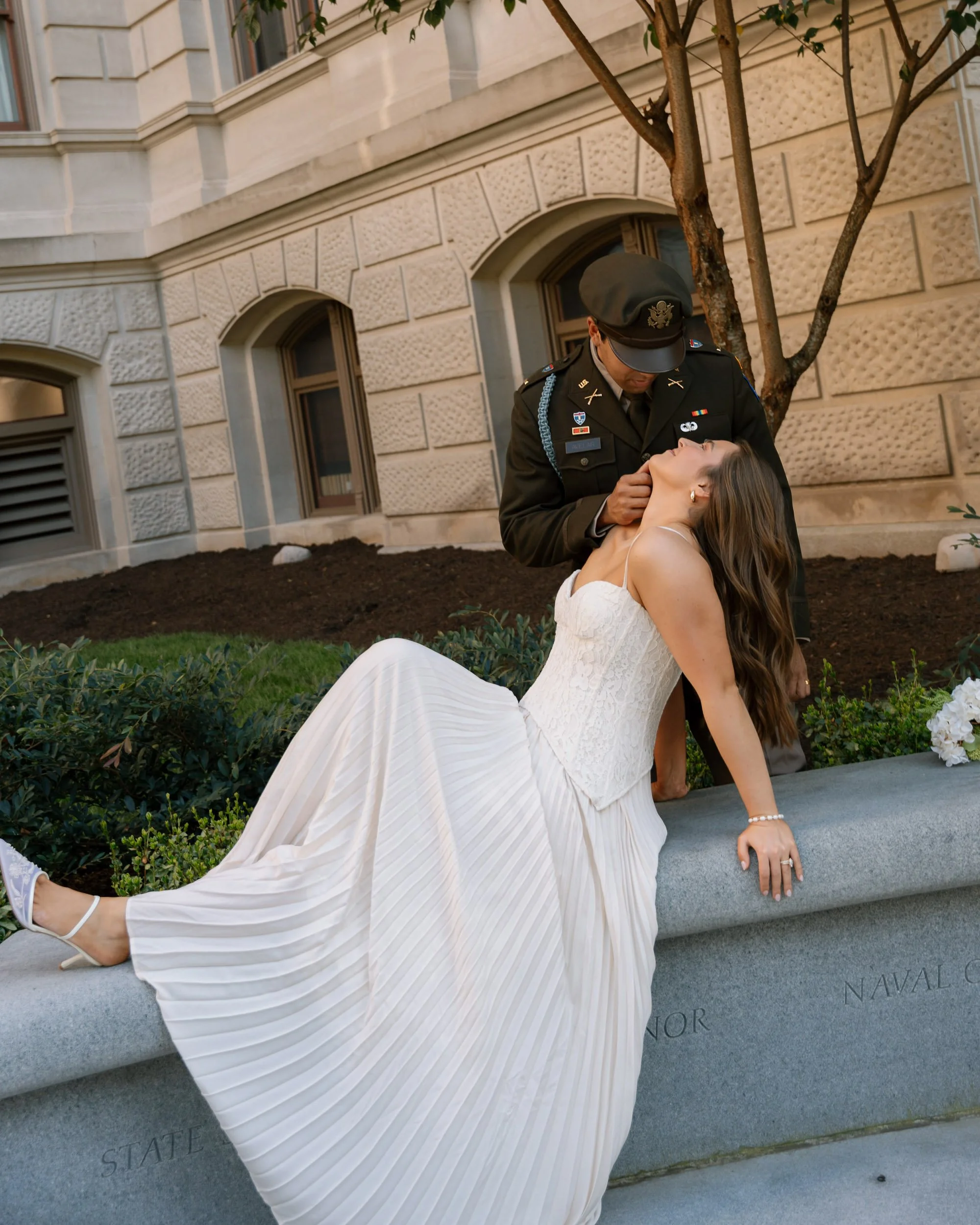 A woman in a white dress with pleated skirt reclines on a stone ledge, while a man in uniform stands beside her, holding her chin during a theatrical or staged moment outside a government building.