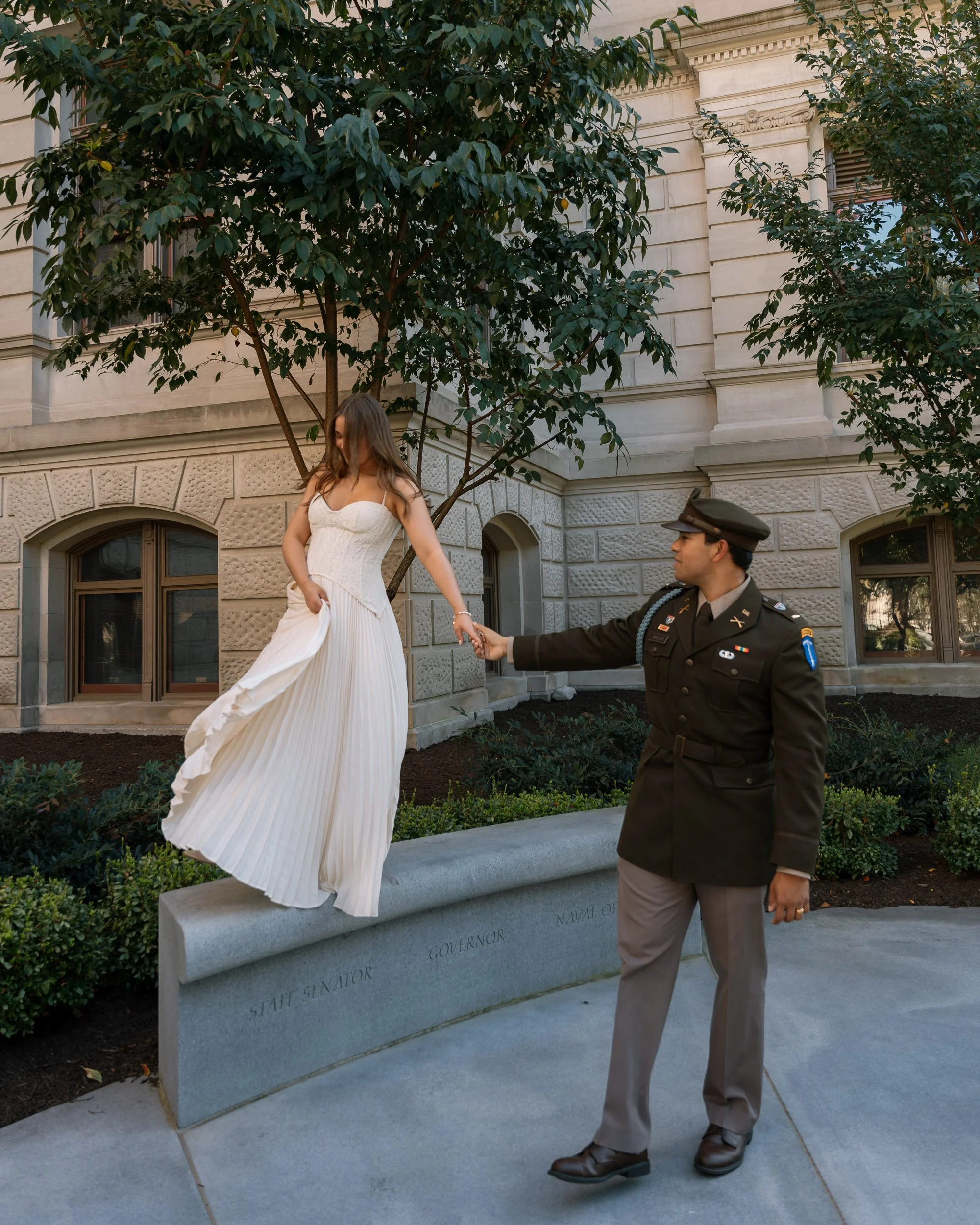 A woman in a white dress standing on a stone bench, holding hands with a man in a military uniform in front of a historic building with trees and windows in the background.