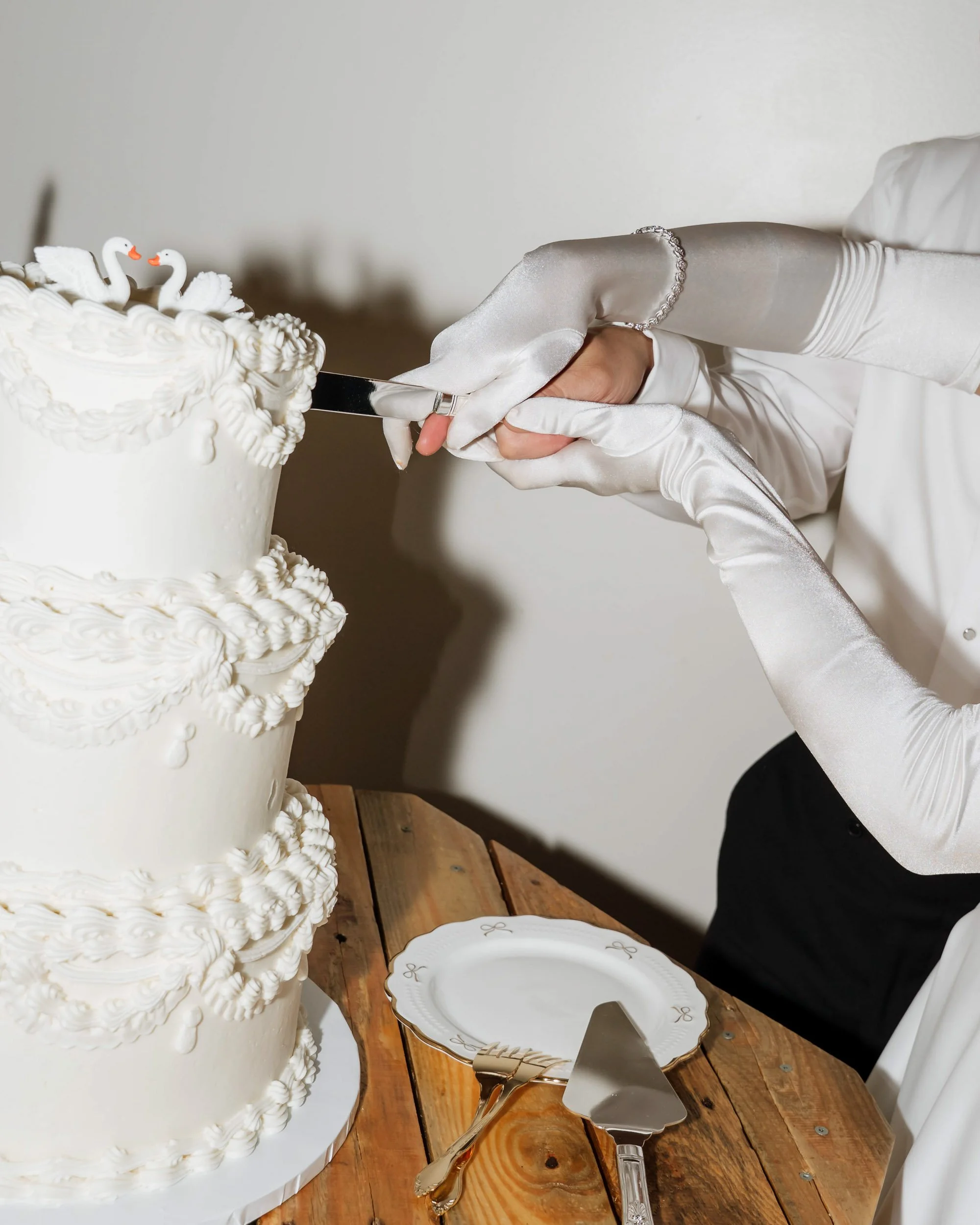 Close-up of someone in white gloves cutting a white, multi-tiered wedding cake with swan decorations on top. The cake is on a wooden table with a gold and white cake server and a white plate nearby.