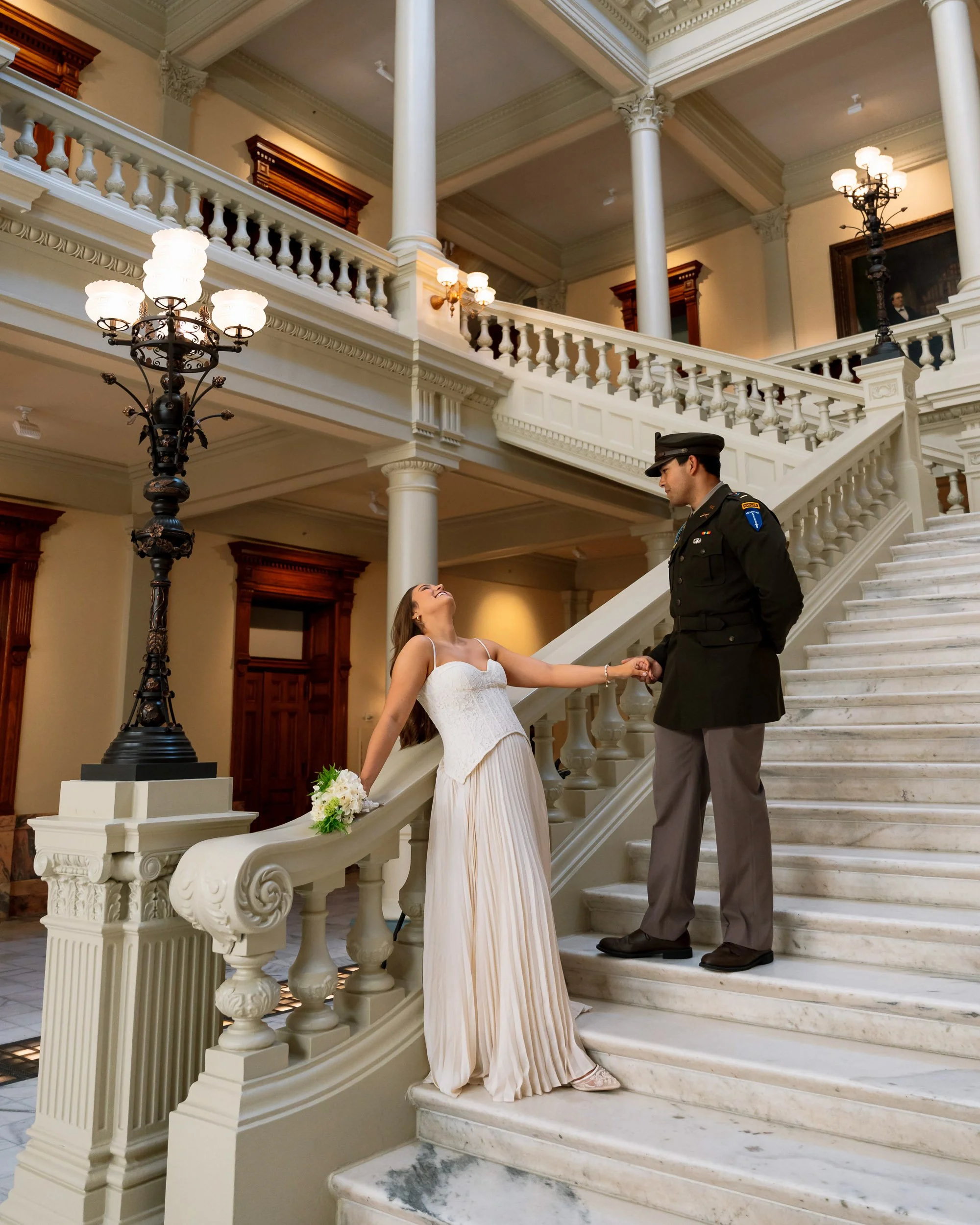 A woman in a white wedding dress leaning back and smiling, holding hands with a man in a military police uniform on a grand staircase inside a historic building.