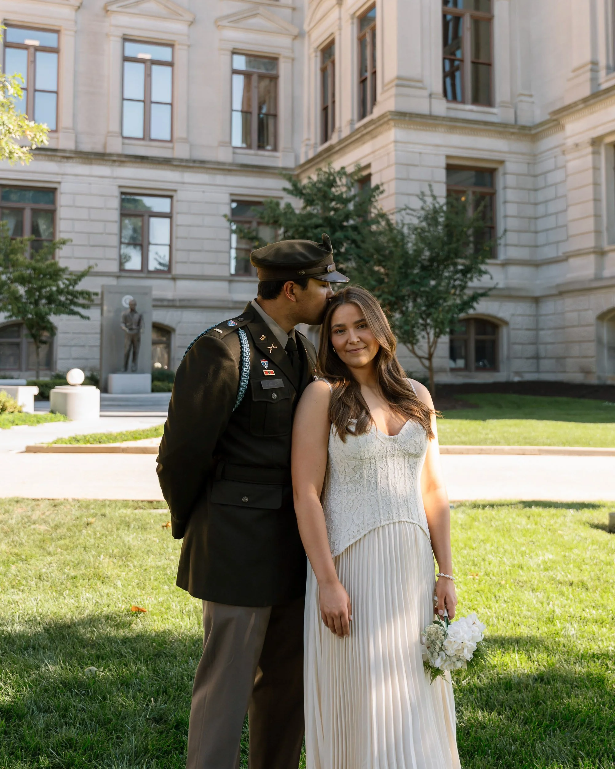 A groom in a military uniform kissing a bride in a white wedding dress holding a bouquet of white flowers, standing on a lawn in front of a large, historic building with multiple windows and a statue.