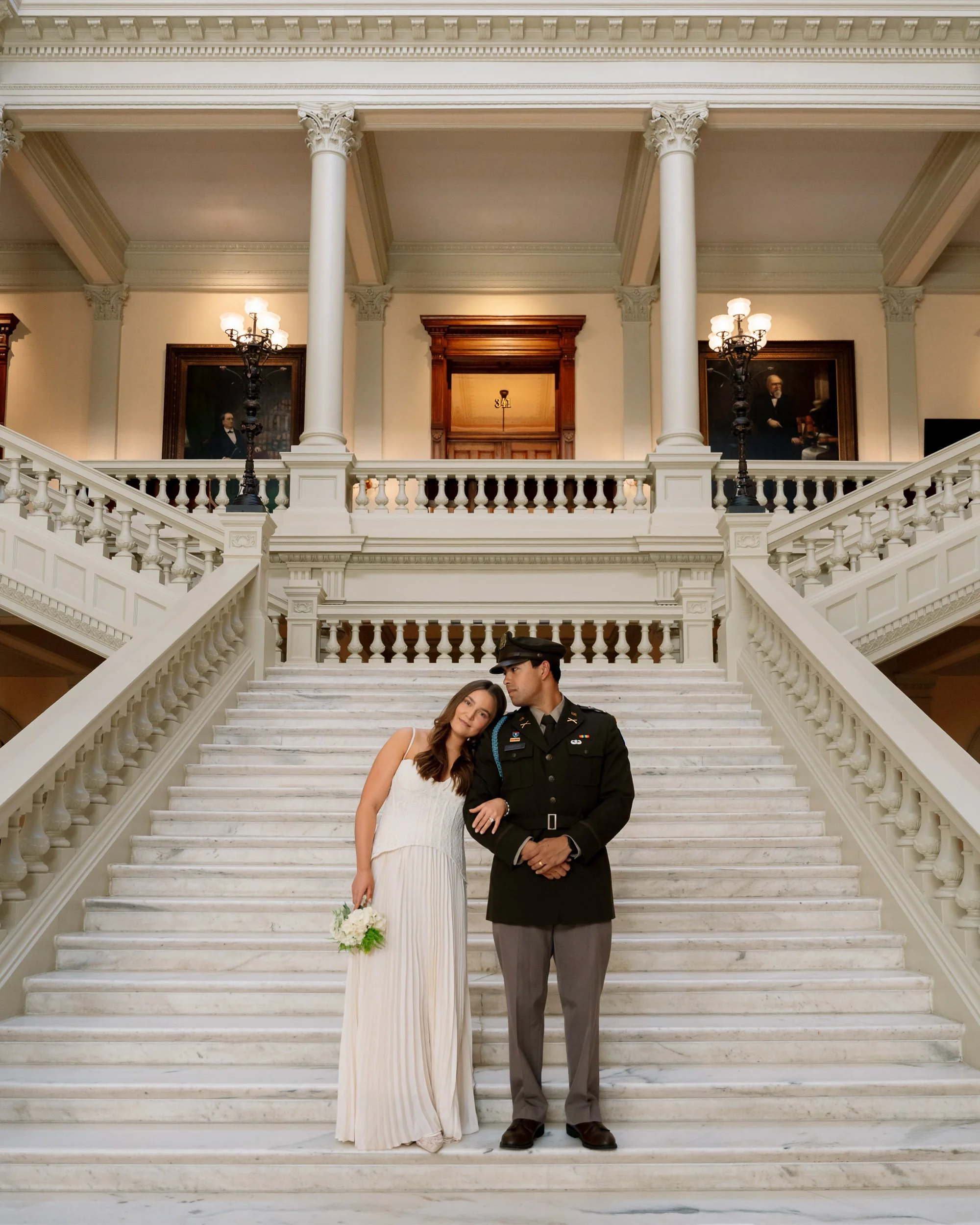 A woman in a white dress holding a bouquet stands next to a man in military uniform on a grand marble staircase inside a large, elegant building with ornate railings, columns, paintings, and chandeliers.