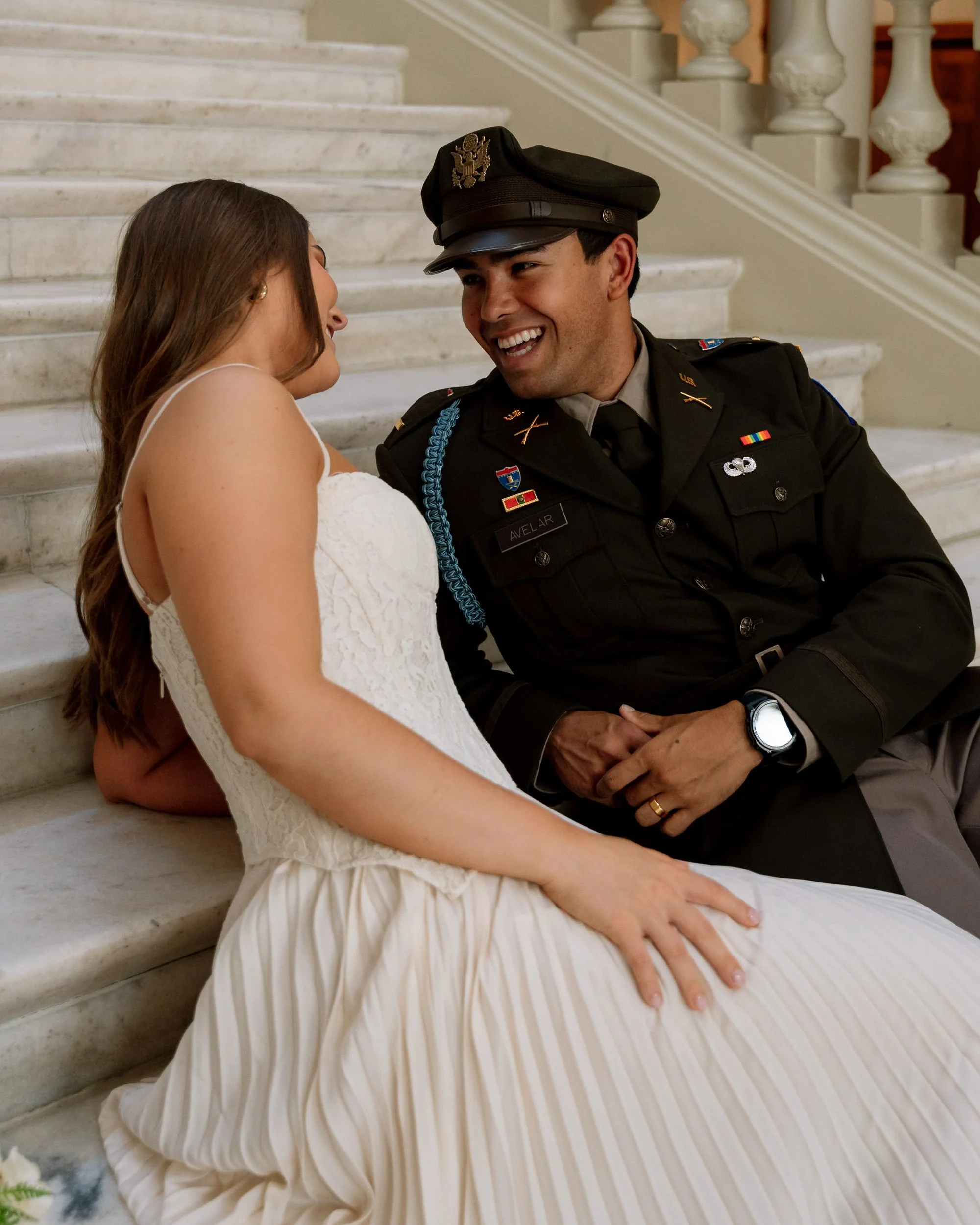 A woman in a white dress and a man in a U.S. military uniform sitting on marble stairs, smiling and looking at each other.