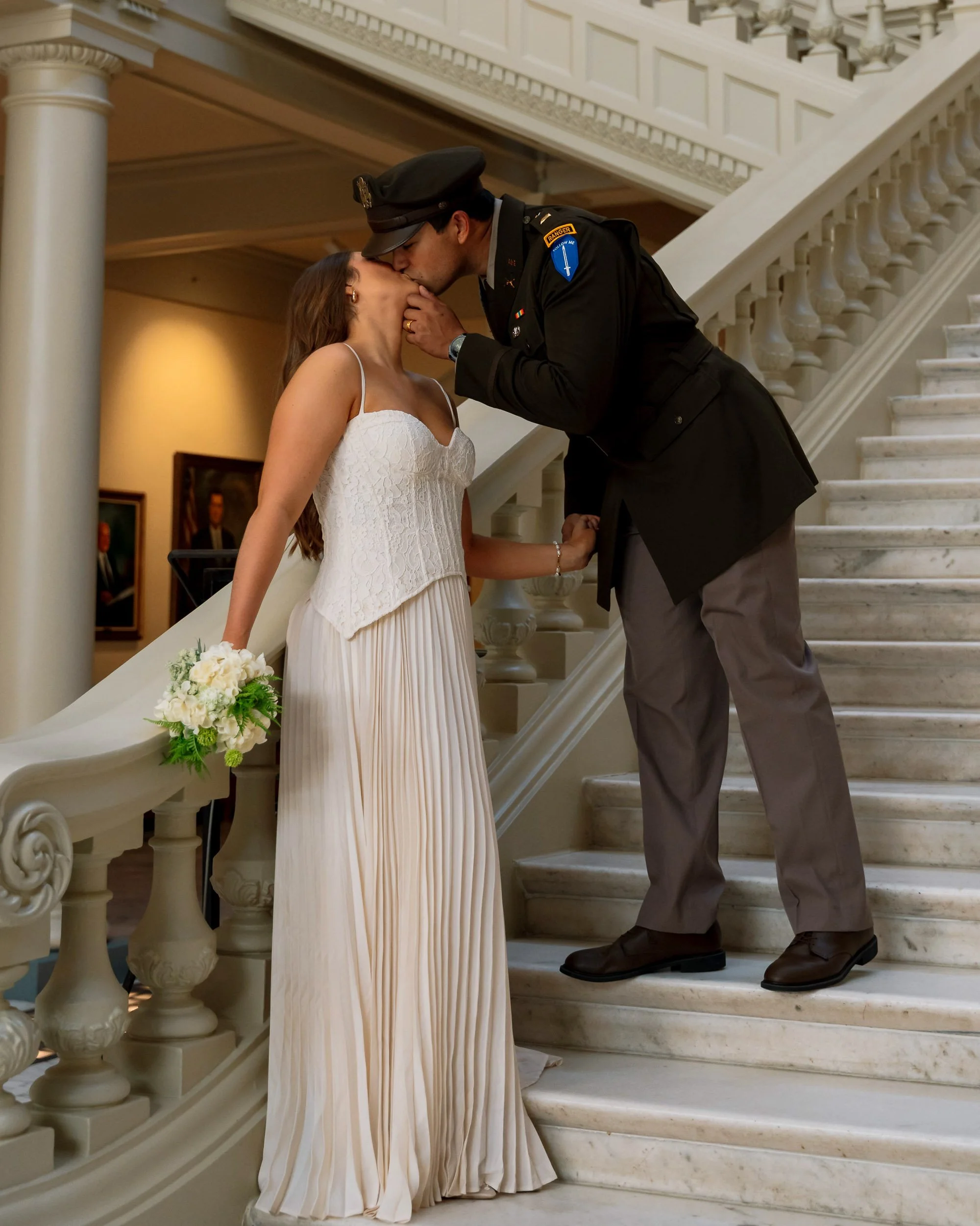 A couple dressed in wedding attire share a kiss on a marble staircase, with the officiant's hand gently holding the woman's face.