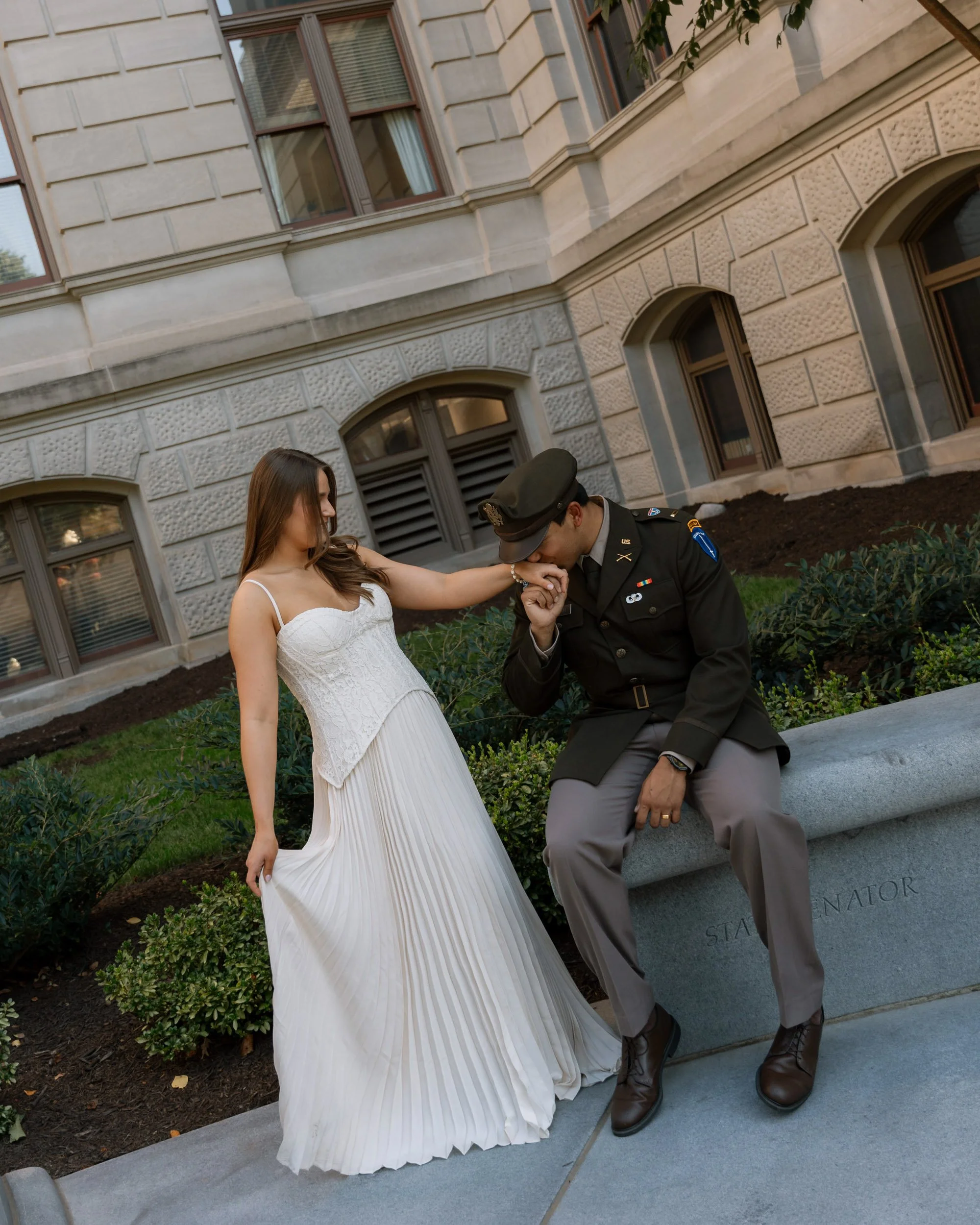 A woman in a white dress kneeling and holding hands with a man in a military uniform sitting on a bench outside a stone building, during daylight.