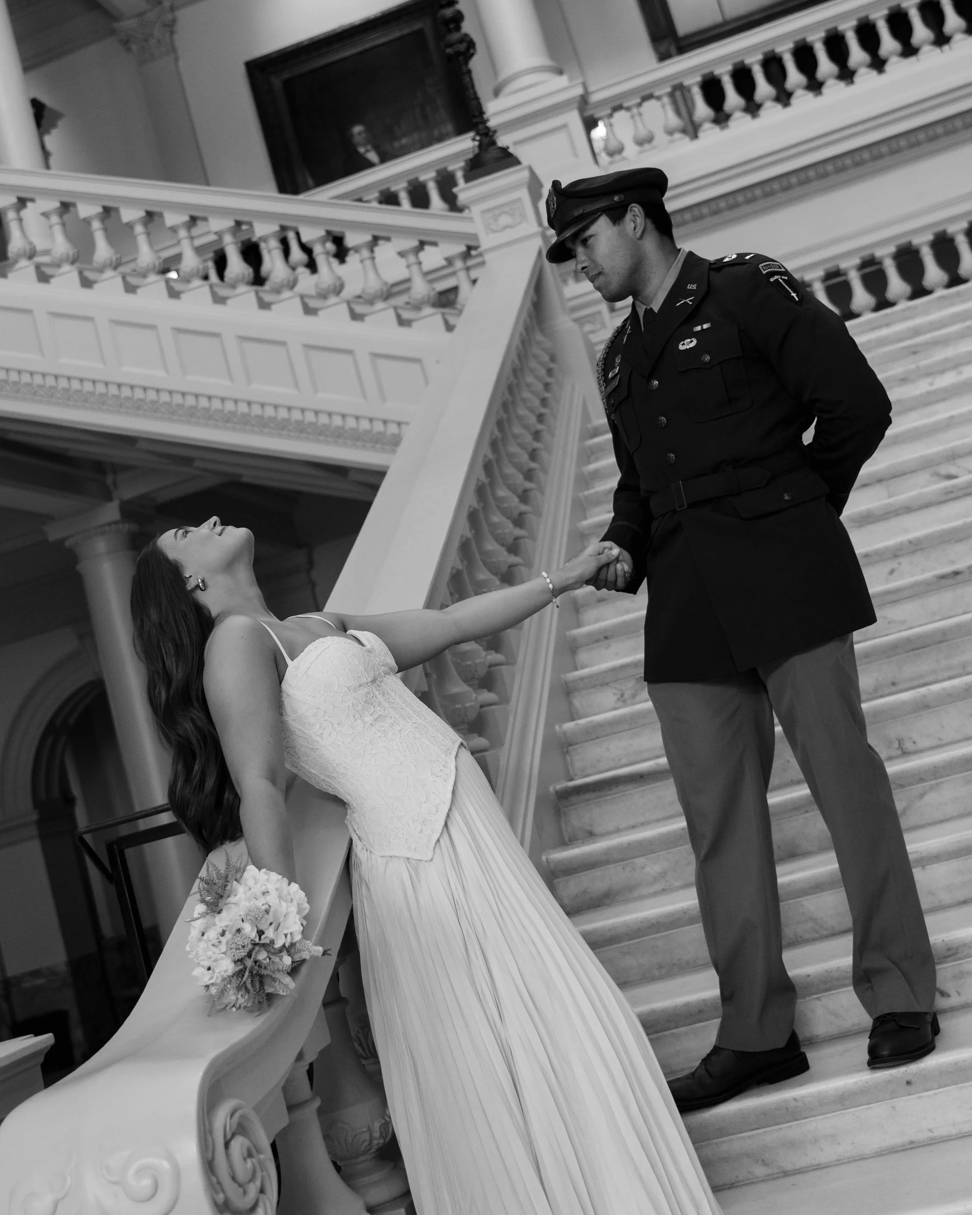 A black and white photo of a woman in a wedding dress holding a bouquet, leaning back and reaching out to a man in a military uniform, standing on a grand staircase inside a large ornate building.