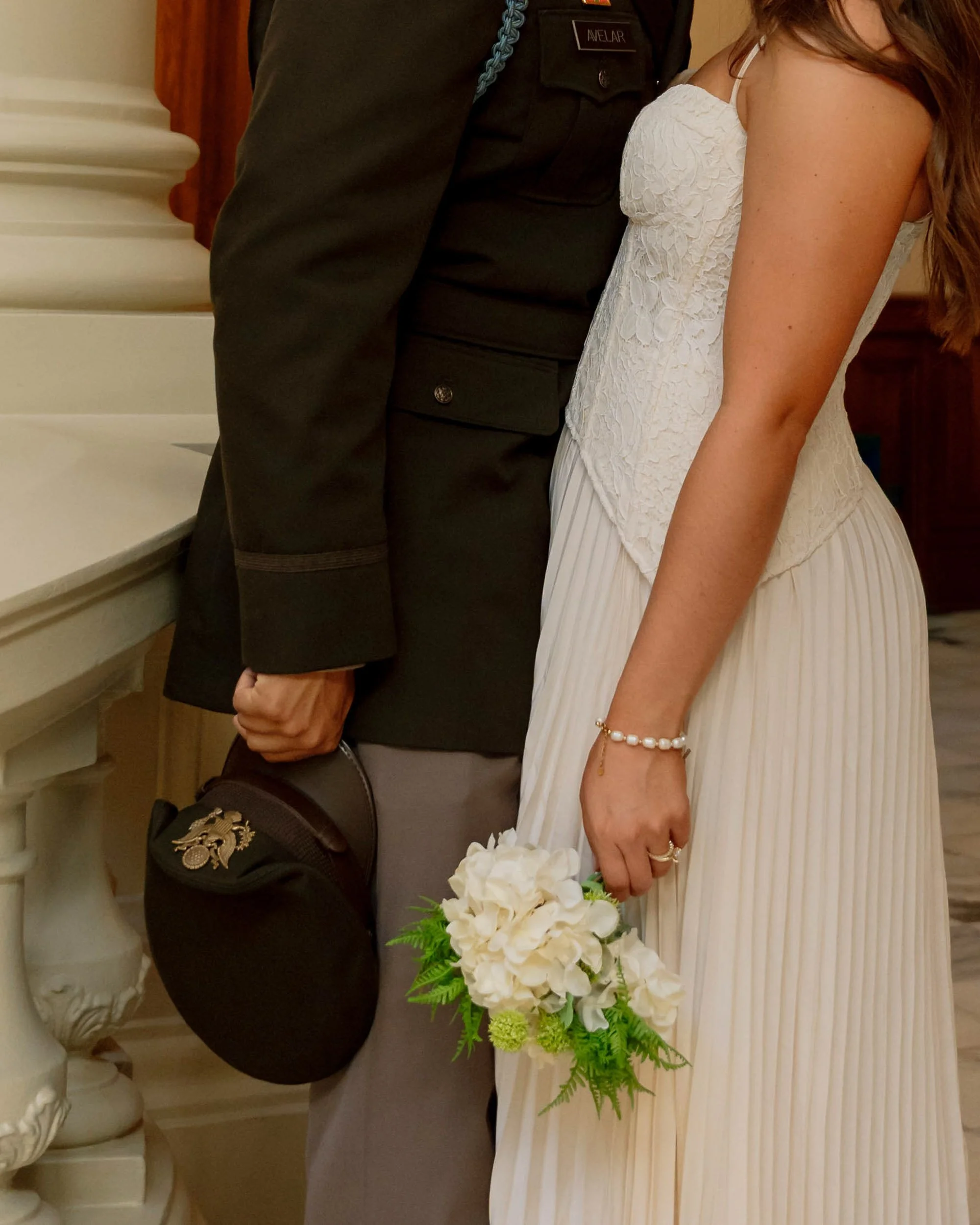 A couple dressed in formal attire, with the woman holding a bouquet of white flowers and the man holding a hat with a military insignia, standing close together in an indoor setting.