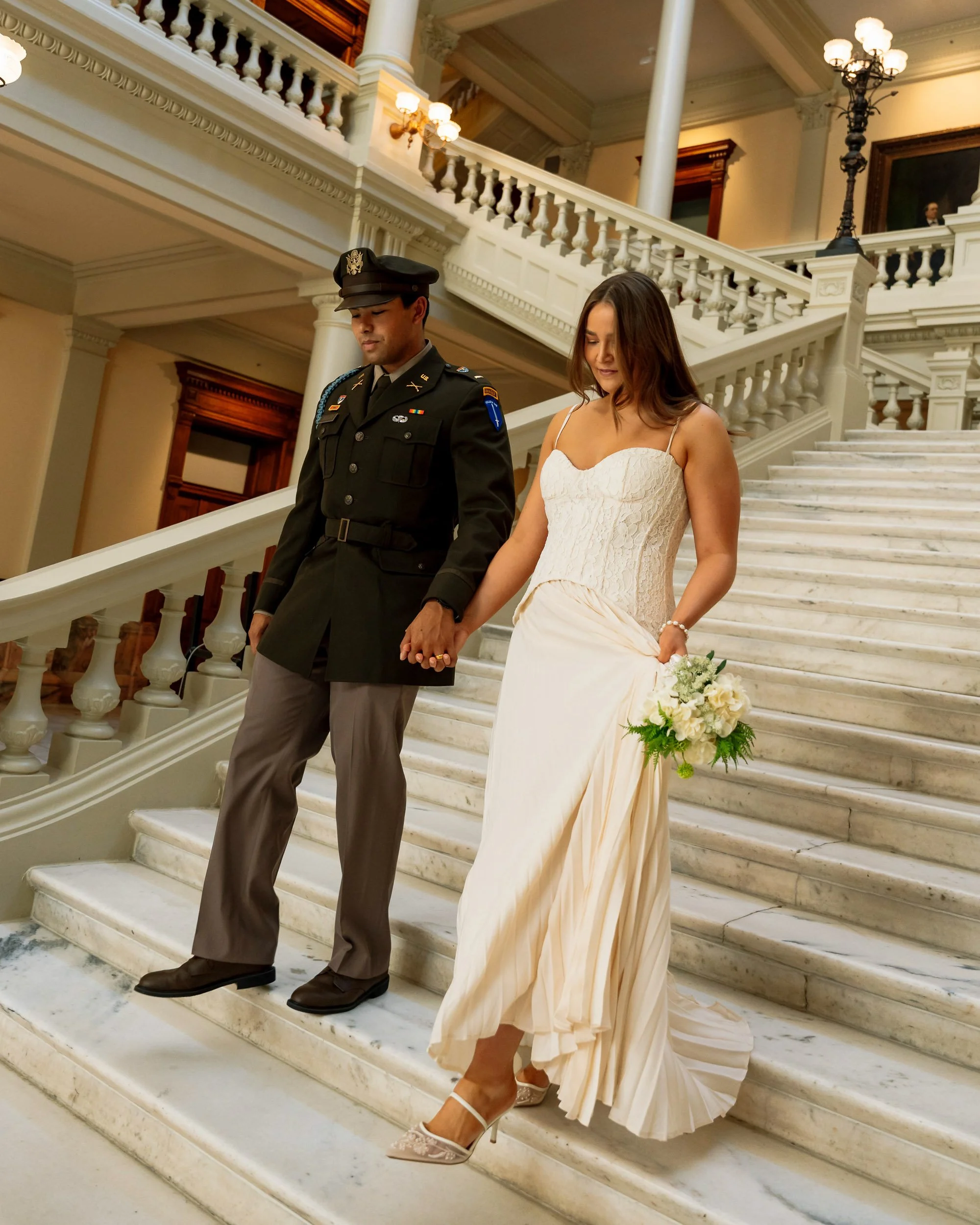 A bride and groom holding hands descending marble stairs in a grand, historic building. The bride wears a white strapless wedding dress and heels, holding a bouquet. The groom is dressed in a military uniform. The setting includes ornate light fixtur