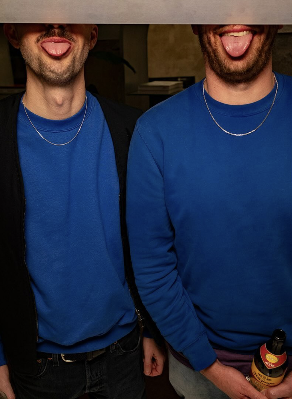 Zwei junge Männer tragen blaue Sweatshirts und ziehen die Zungen heraus, stehen nahe beieinander, einer hält eine Flasche Bier, im Hintergrund ein Regal mit Büchern.