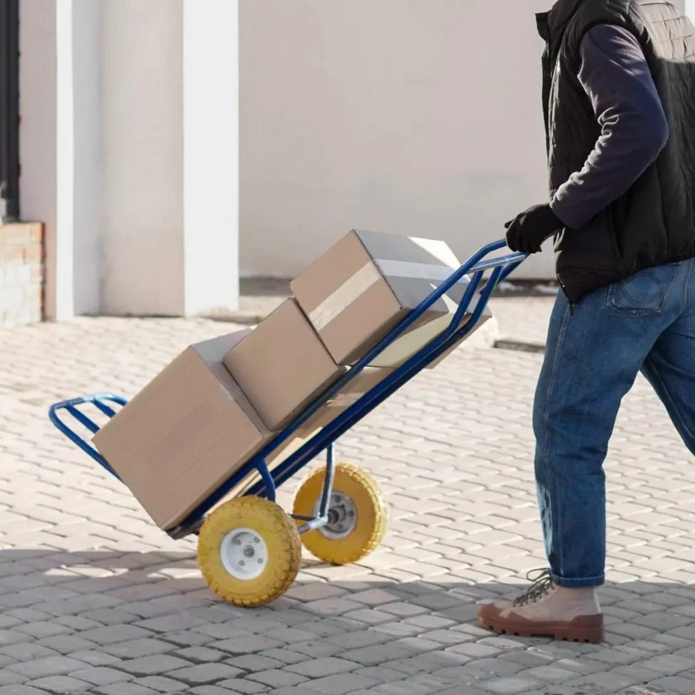 Person wearing boots, jeans, black gloves, and a black jacket pushing a blue hand truck with three cardboard boxes on cobblestone ground outside.