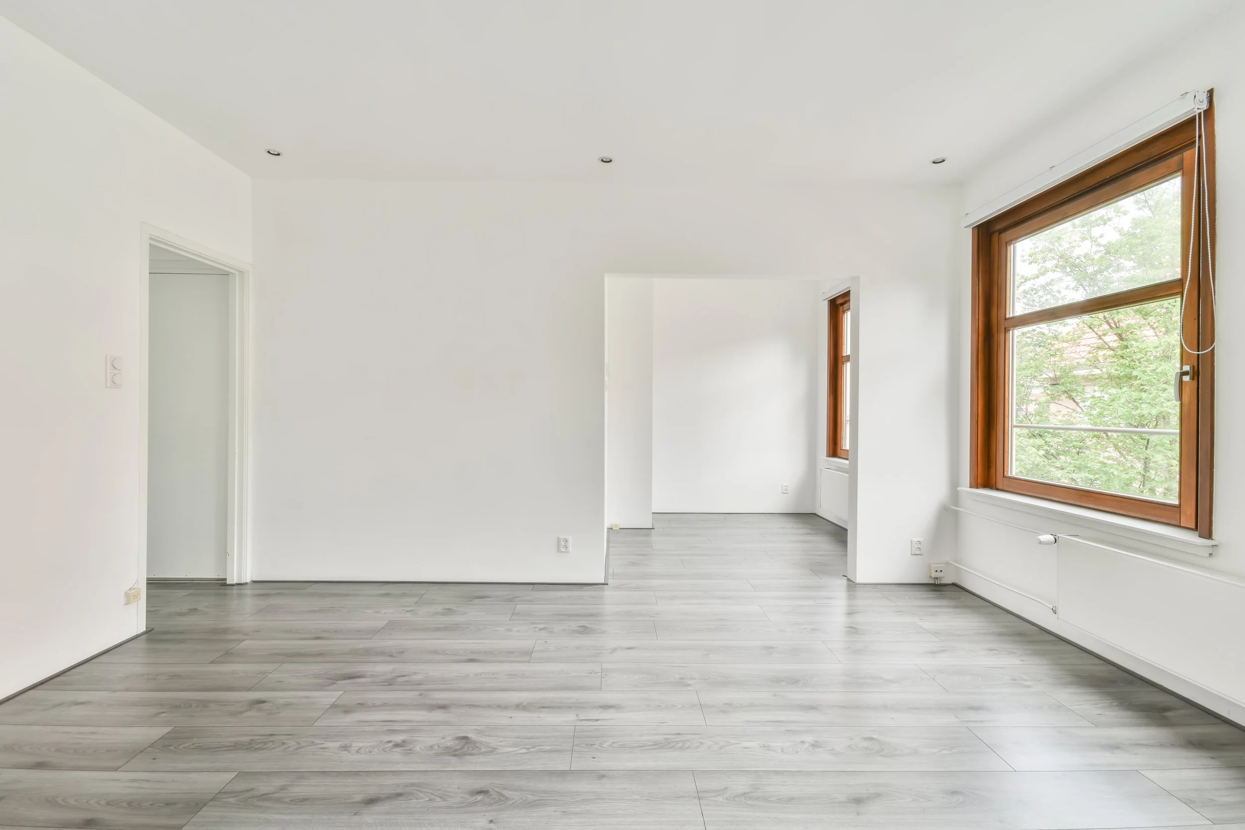 Empty room with white walls, gray wood flooring, large windows with wooden frames, and natural light.