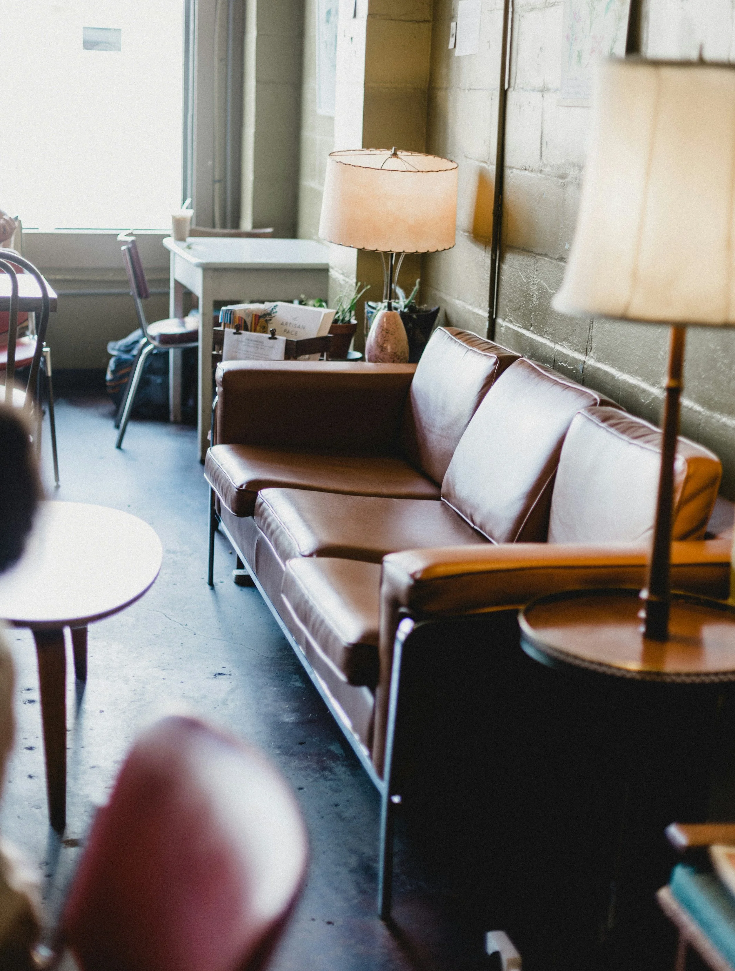 Interior of a cozy café with a brown leather sofa, two standing lamps with beige shades, a small round table, and a window letting in natural light.