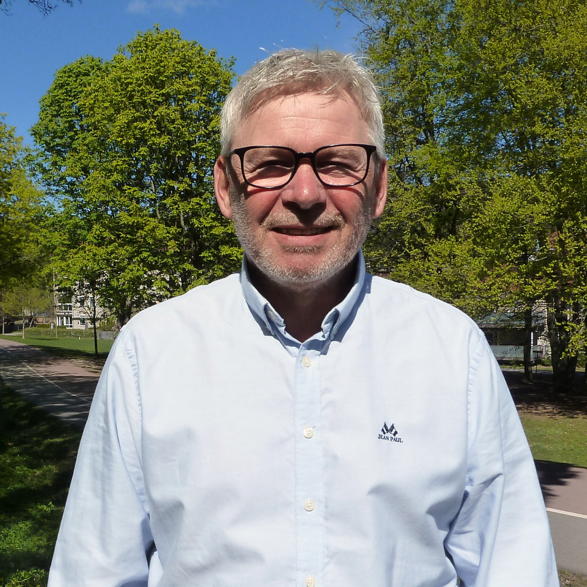 En mann med grått hår og skjegg, smilende, iført en blå genser og skjorte, står foran en lys vegg.