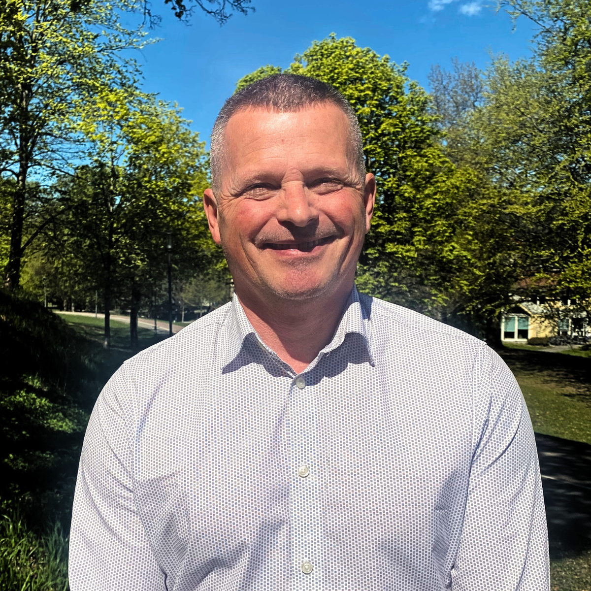 Portrettbilde av en person med kort brunt hår, blå øyne, smil, og rød-hvit rutete skjorte, stående mot en lysegrå bakgrunn.