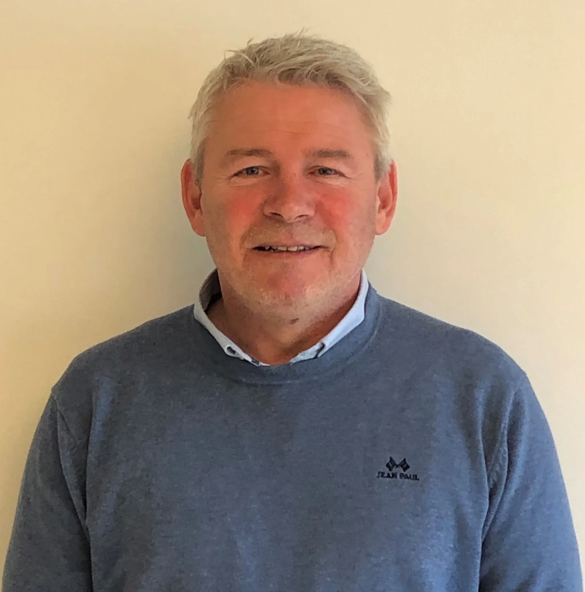 En kledd mann med grått hår, iført en blå genser med logo.