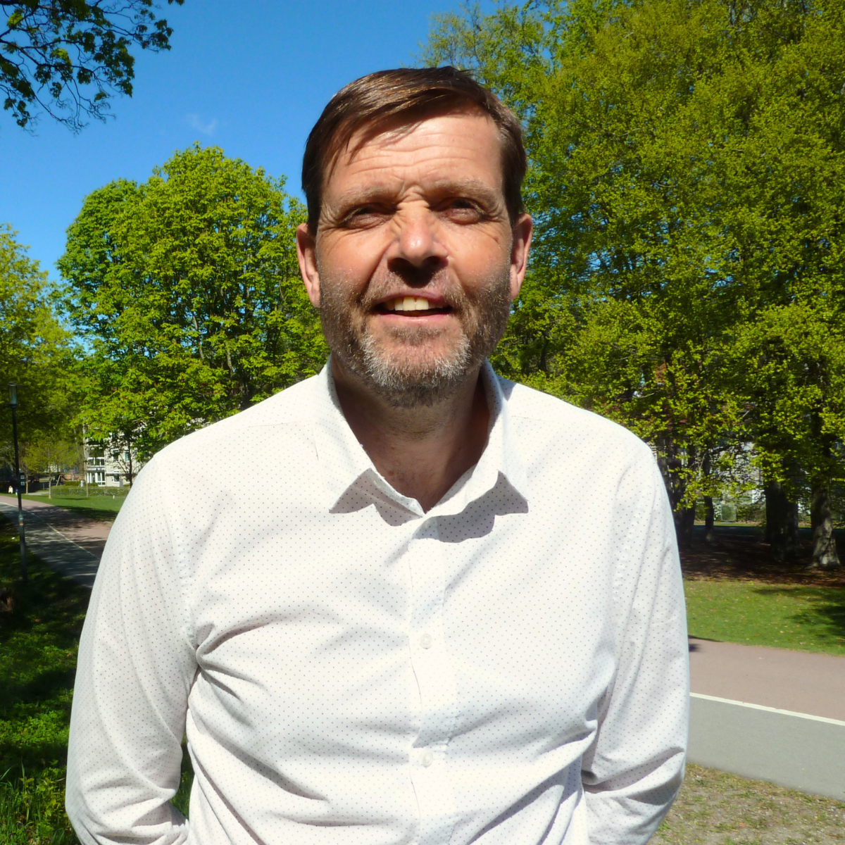 Portrett av en mann med mørkt hår, iført blå skjorte og mørk genser, stående foran en lys vegg.