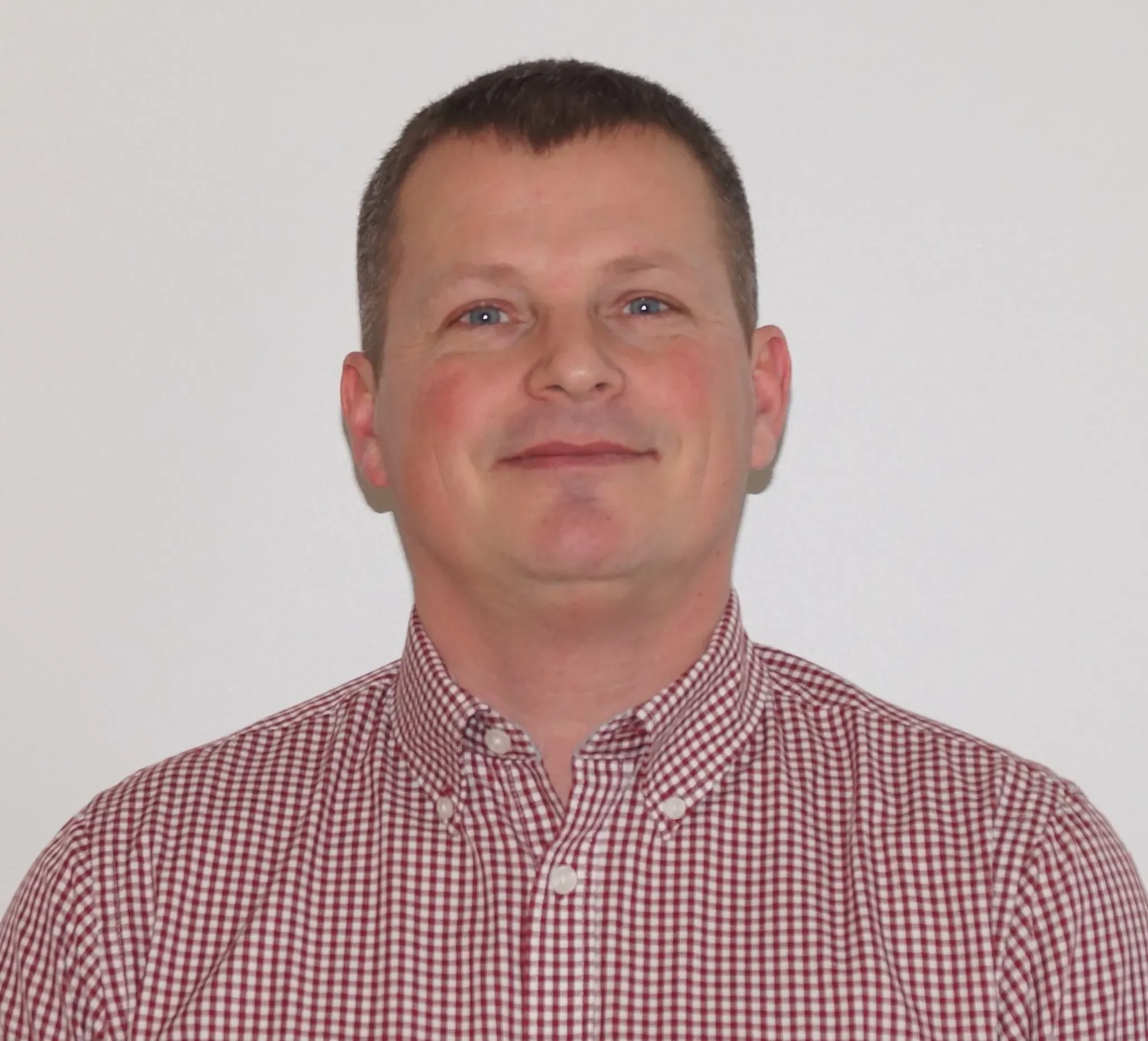 Portrett av en mann med kort hår, blå øyne, iført rød og hvit rutete skjorte, står foran en lys bakgrunn.