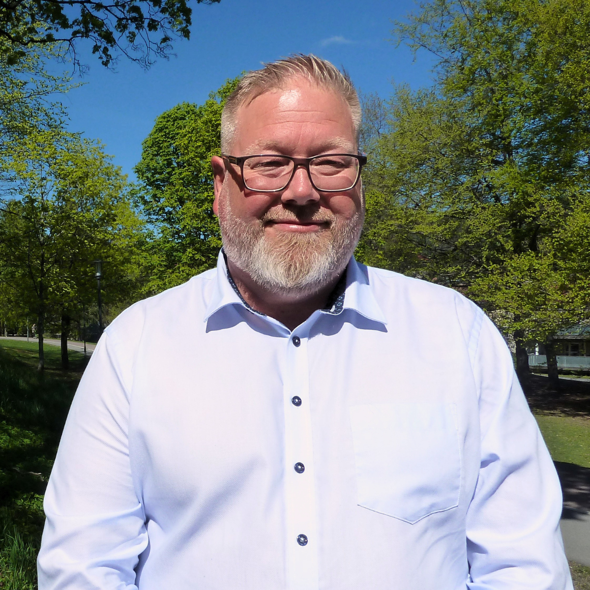 Et foto av en smiling mann med brunt, kort hår og skjegg, iført en blå rutete skjorte med logo på brystet, mot en hvit bakgrunn.