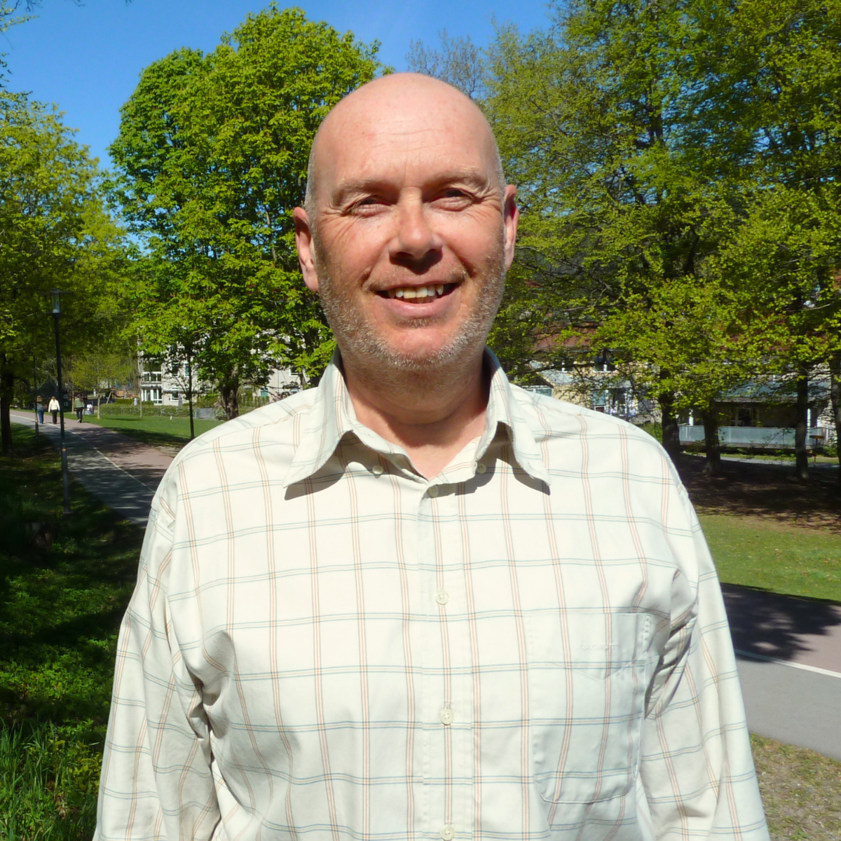Portrett av en mann med skjegg og skjorte med liten rutemønster, smilende mot kameraet.