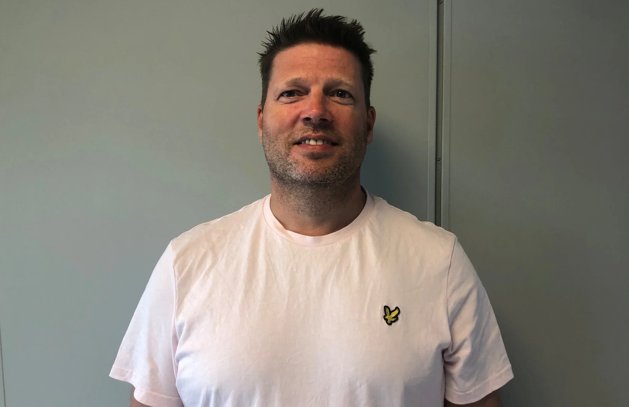 En mann med mørk, spiky hår står foran en lys grå vegg, iført en lys rosa t-skjorte med en liten gul og svart logo på venstre bryst.