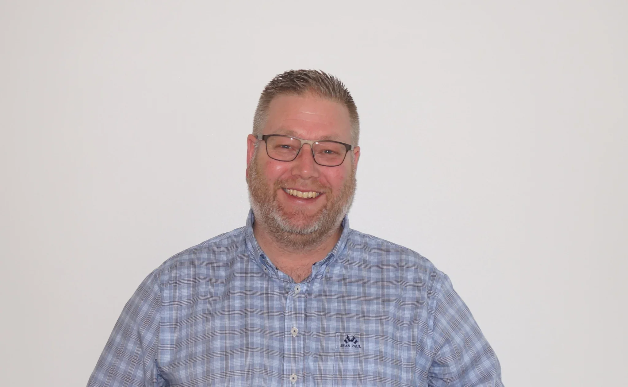 Et smilende man med briller, skjorte og raskt hårposisjon foran en hvit vegg.