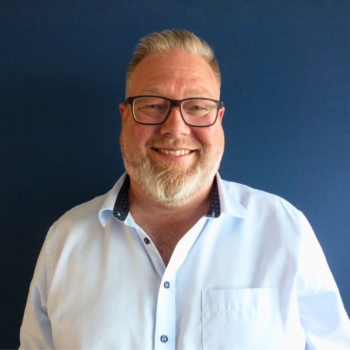 Et smilende man med briller, skjorte og raskt hårposisjon foran en hvit vegg.