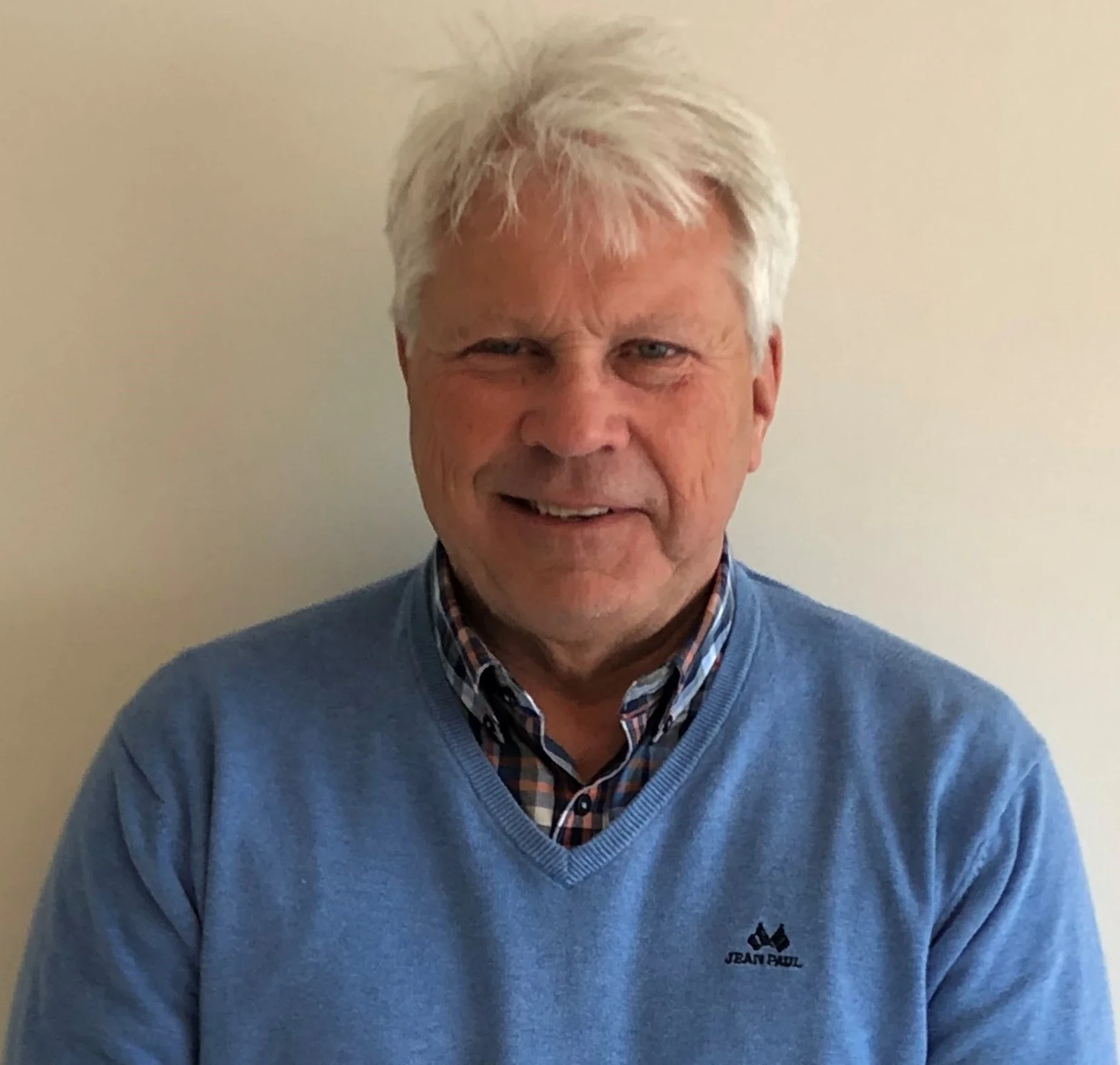 En eldre mann med blond, litt grått hår, smilende, iført blå genser og skjorte, står foran et nøytralt vegg.