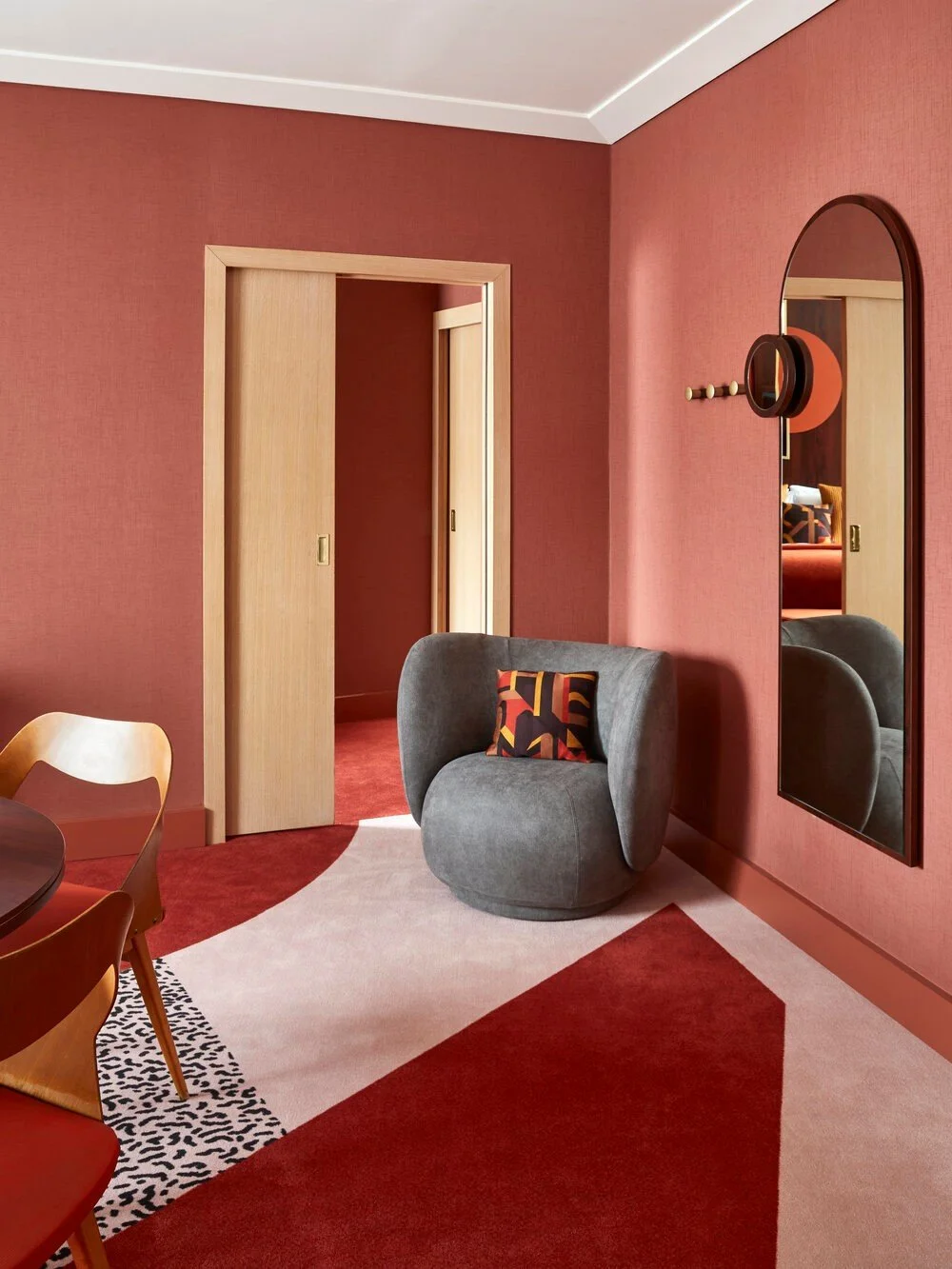 Interior of a room with a red and beige color scheme, featuring a gray modern chair with a decorative pillow, red and beige geometric carpet, and a round wood-framed mirror on the wall. Light wood panel sliding doors are partially open.
