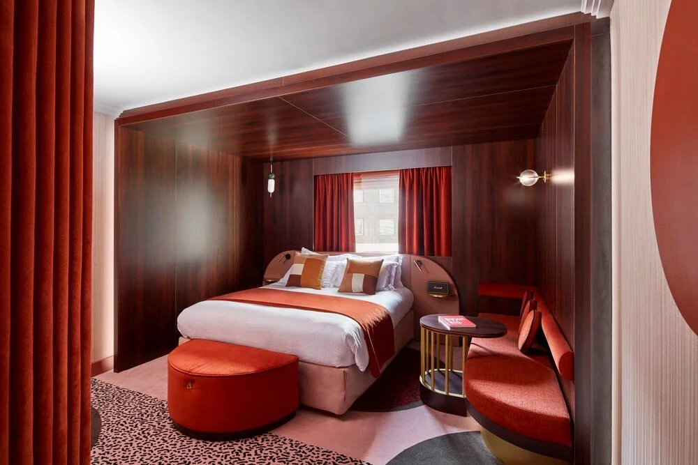 Modern hotel room with a double bed, brown and red decor, round ottoman, small table, and cushioned bench seating.
