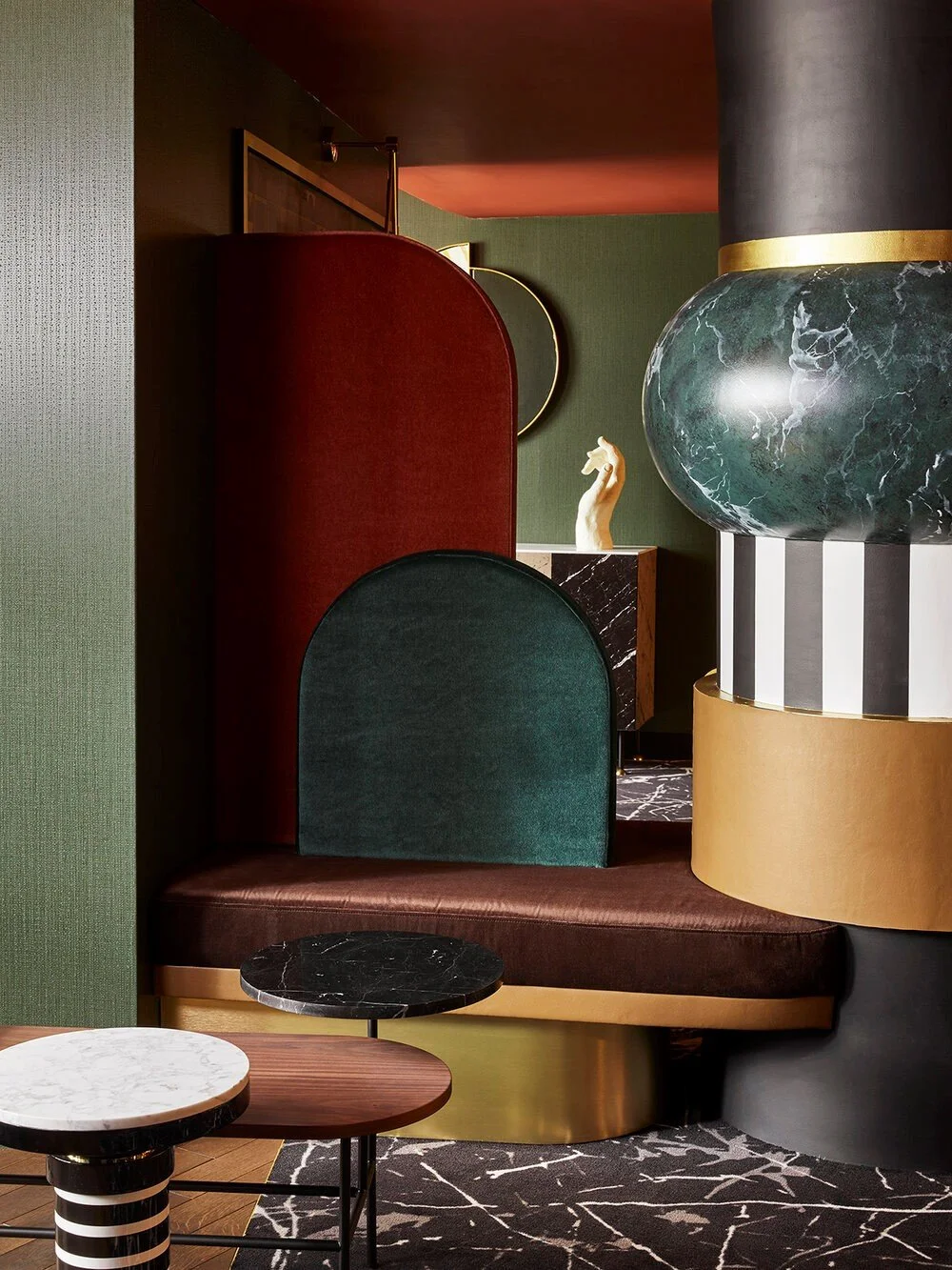 Modern interior with geometric shapes, featuring a bench with curved velvet backrests in red and green, round marble tables, and bold architectural columns with green marble texture and gold accents.