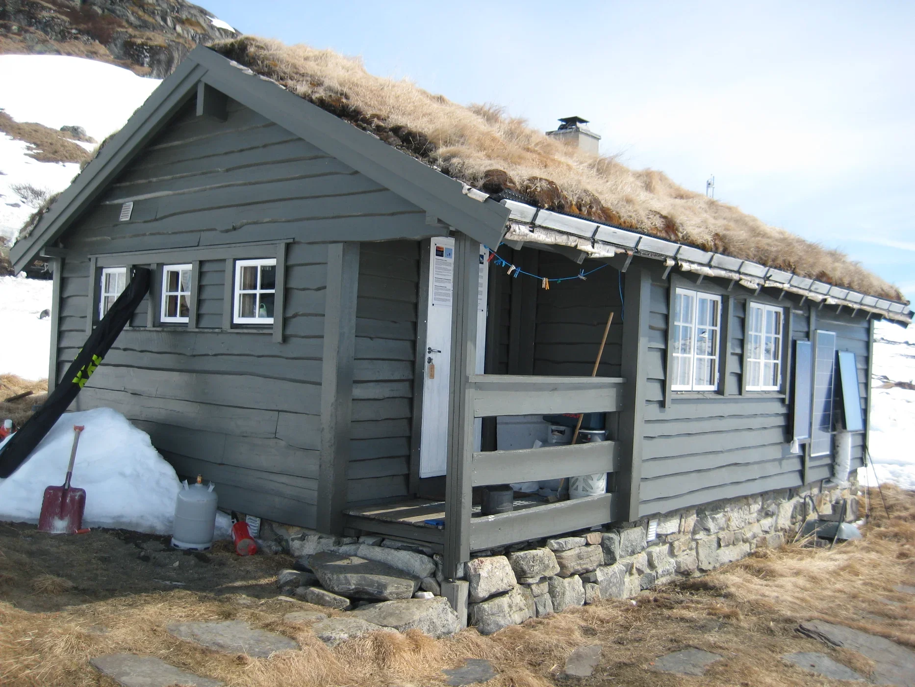 En liten svart trehytte med to små vinduer har en inngang med trapp. Det er snø og noen steiner rundt hytten, og graset rundt er brunt. Himmel er delvis overskyet.
