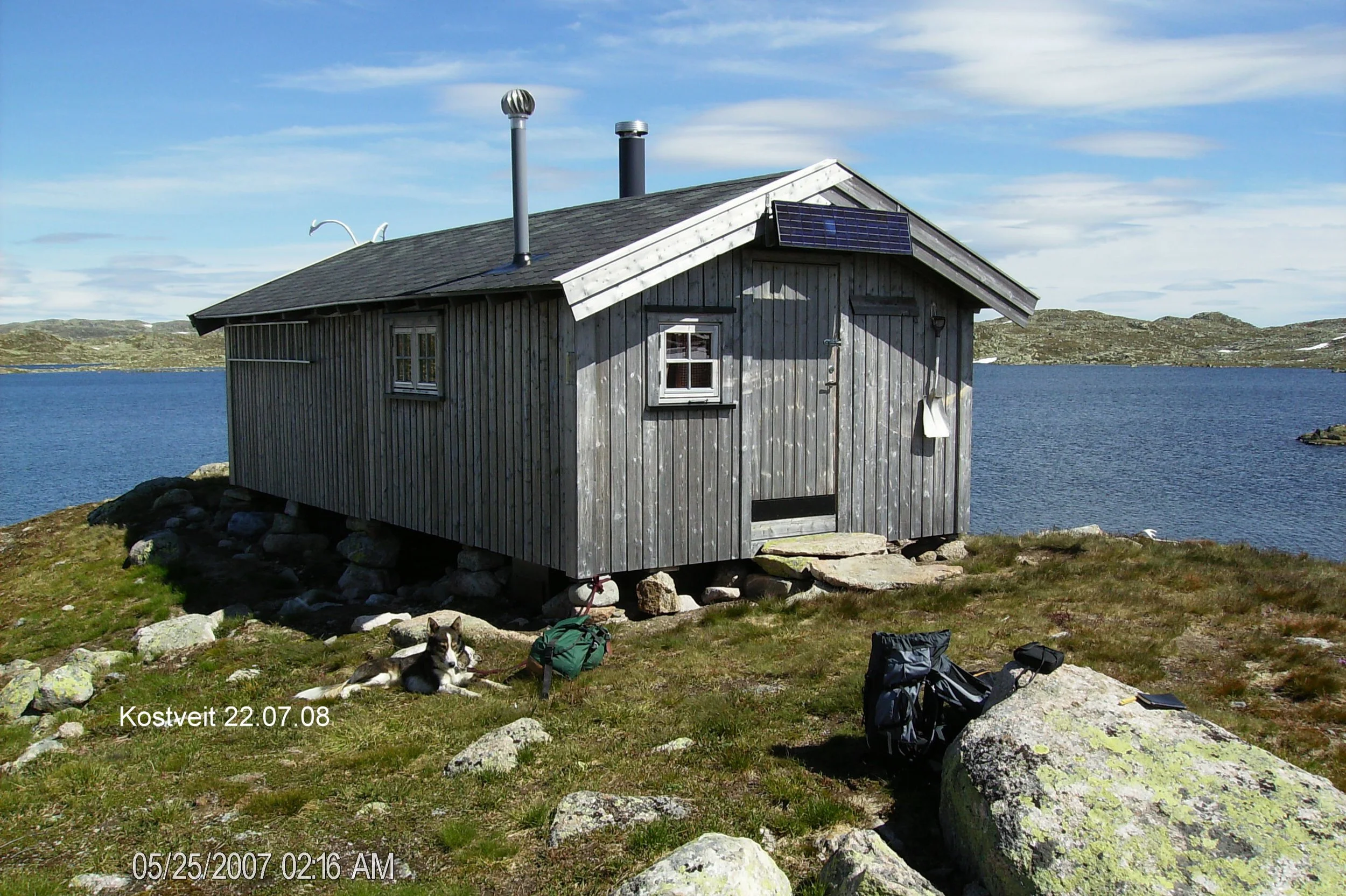 En liten hytte på fjellet ved vann, omgitt av steiner og gress. To hunder ligger på bakken utenfor hytta, og det er ryggsekker og utstyr på bakken. Hytta er avtrekk med trepanel, og det er sol og klar himmel.