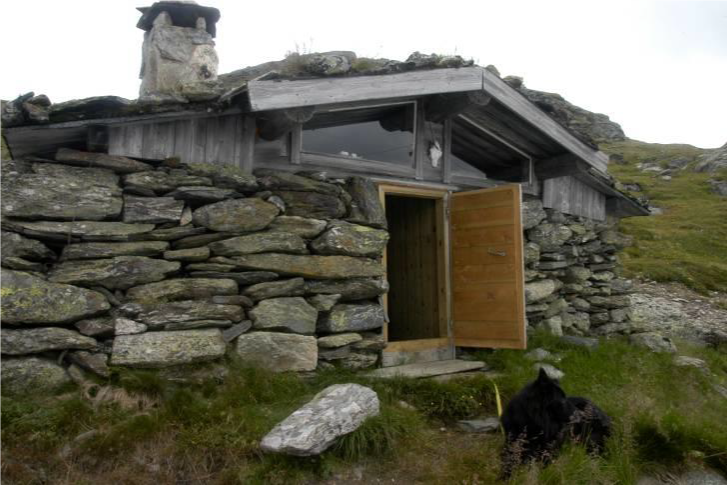 En små stein hytte med åpent døra og en hund som ligger på bakken foran den, omgitt av fjell og gress.