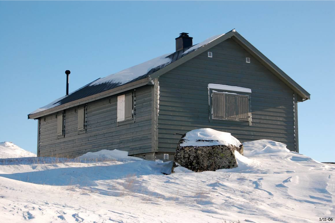 En mørk trehytte med skiftene vinduer, omgitt av snø, under en klar blå himmel.