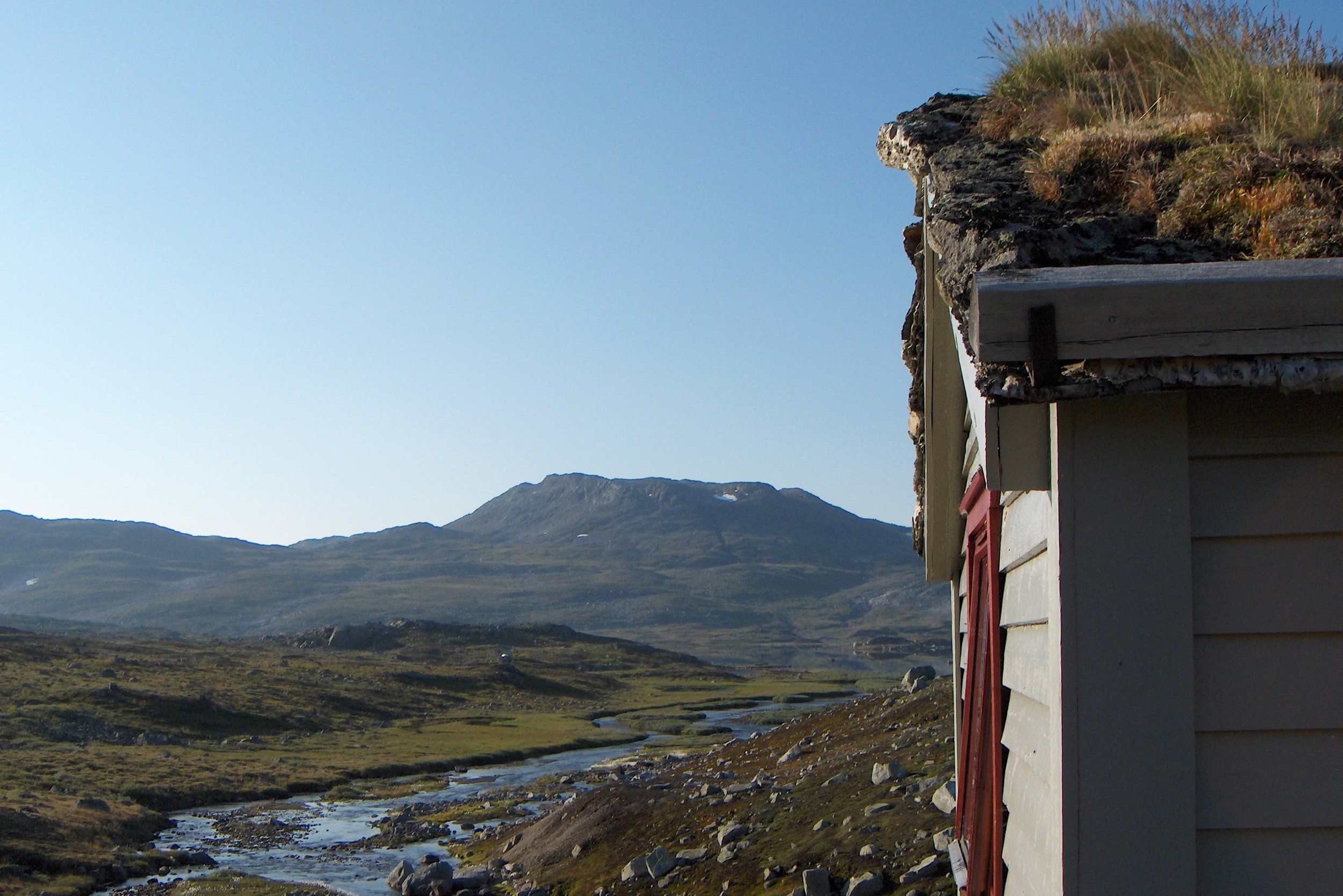 Et lite hus med vegg og tak i fjellandskap, med elv og fjell i bakgrunnen.