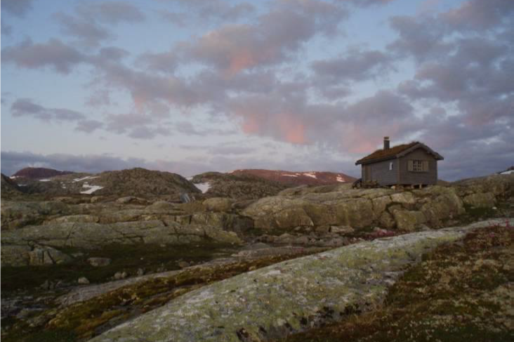En liten hytte på steinete terreng under en skyet himmel ved solnedgang.