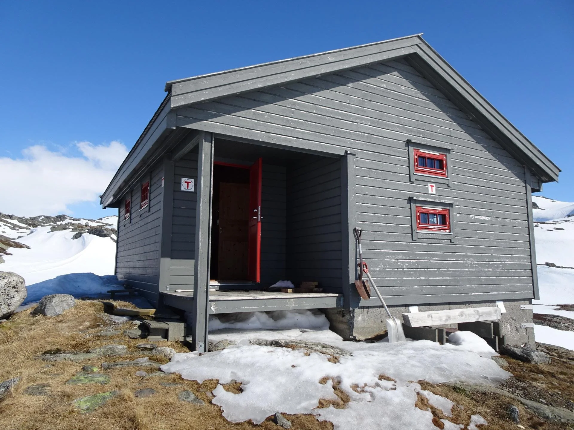 En liten, grå tømret hytte med røde vindusrammer og åpen dør, plassert i snødekt fjelllandskap under en blå himmel.