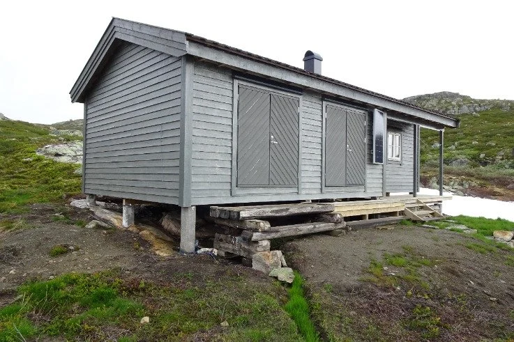 Lite blått hytte med utvendige vegger av tre, hevet på støttebjelker med enkle tre-trinn opp til inngangsdøren, omgitt av fjell og myrlandskap.