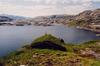 En grønn ås med et lite murbygning på toppen ved en innsjø omgitt av fjell.